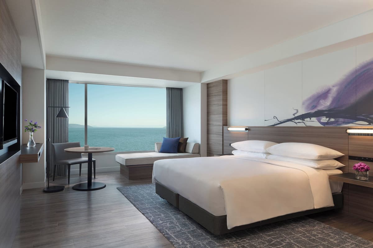 Une chambre d’hôtel moderne dotée d’un grand lit, d’un coin salon avec vue sur la mer, d’une télévision à écran plat et d’un grand tableau abstrait accroché au mur.