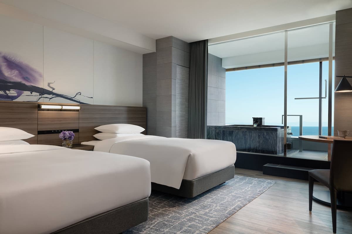 Une chambre d’hôtel moderne avec deux lits, une grande fenêtre donnant sur la mer, un balcon et une petite table avec une lampe.