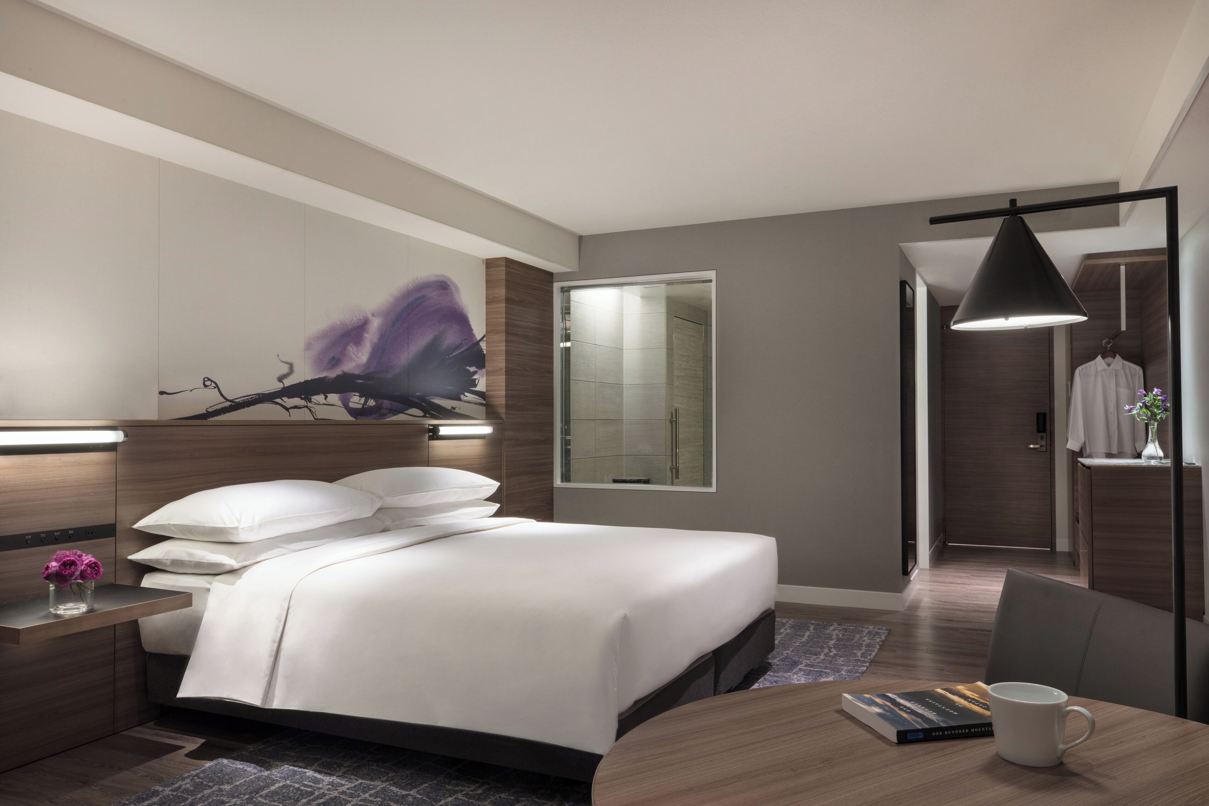 Una habitación de hotel moderna con una cama bien hecha, una mesita de noche con un florero y un ventanal con vistas al baño.