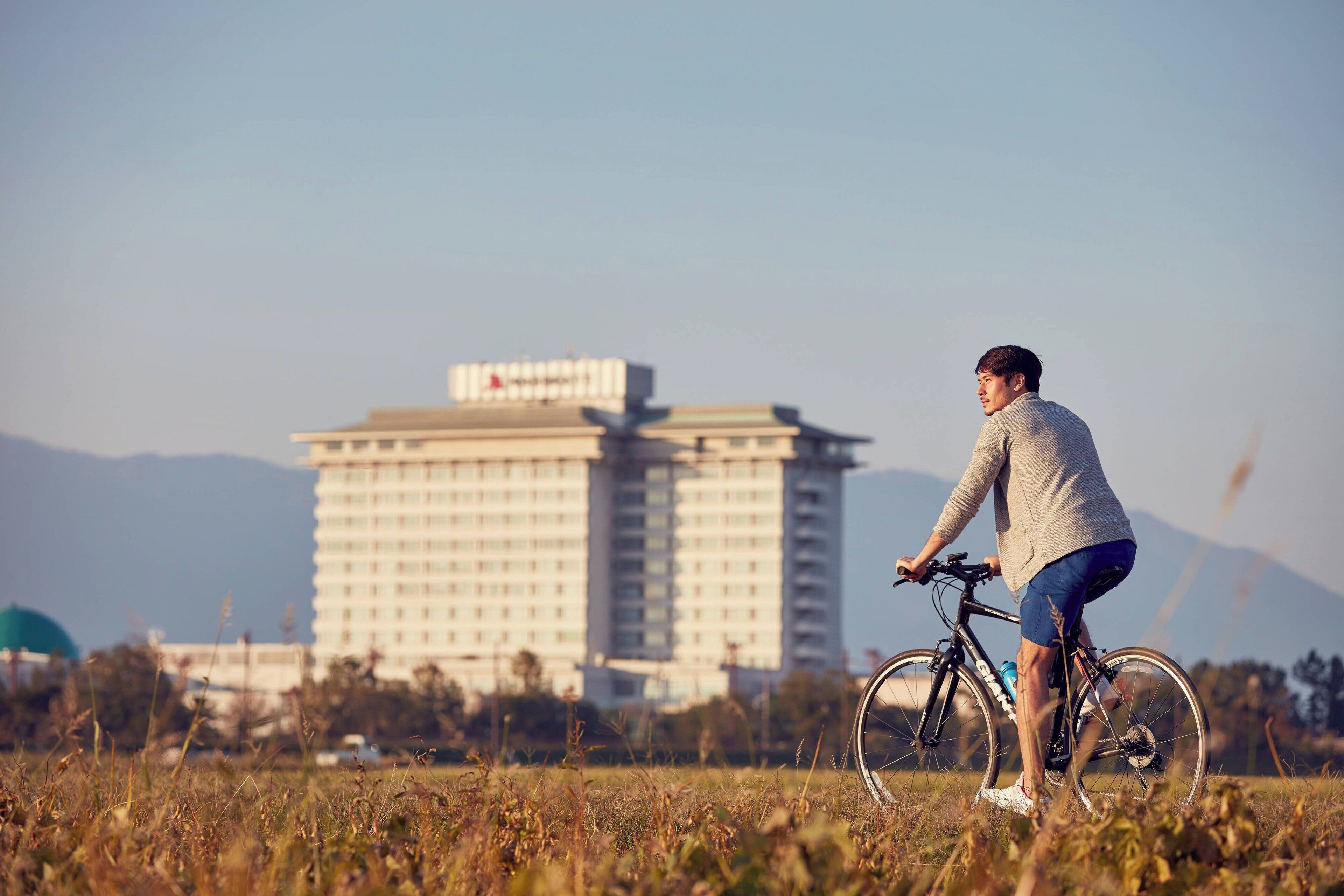 A person riding a bicycle in a grassy field with a large hotel building in the background.