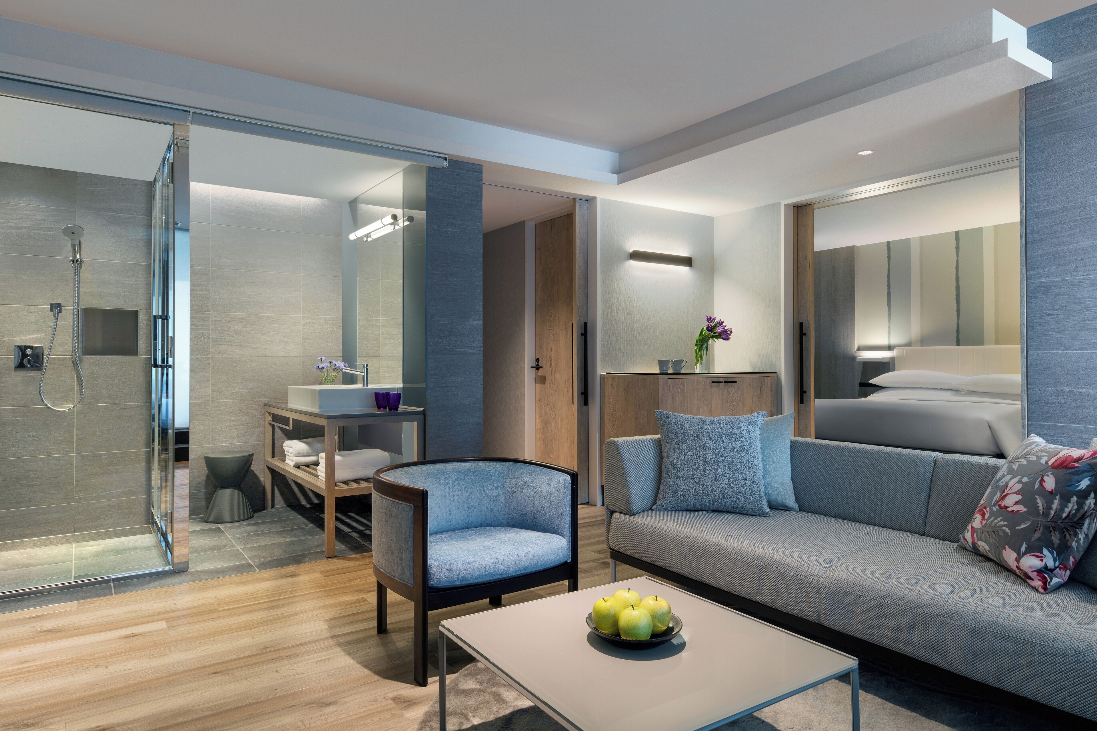 A modern hotel room with a shower, a sofa, a chair, a small table with apples, and a bedside table with a vase.