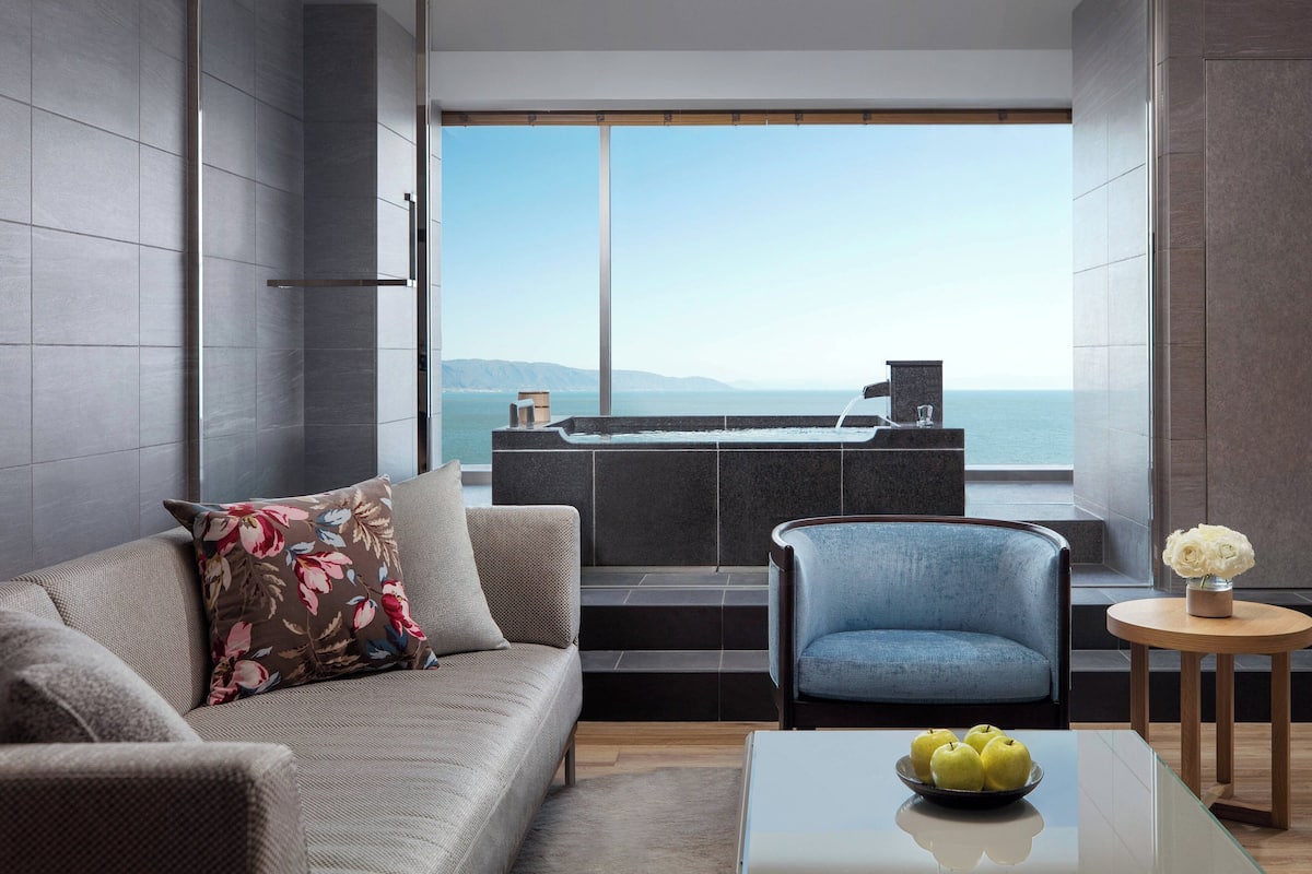 Un salon moderne avec un canapé, un fauteuil bleu et une table basse sur laquelle sont posées des pommes. Une grande fenêtre offre une vue sur la mer et les collines lointaines.
