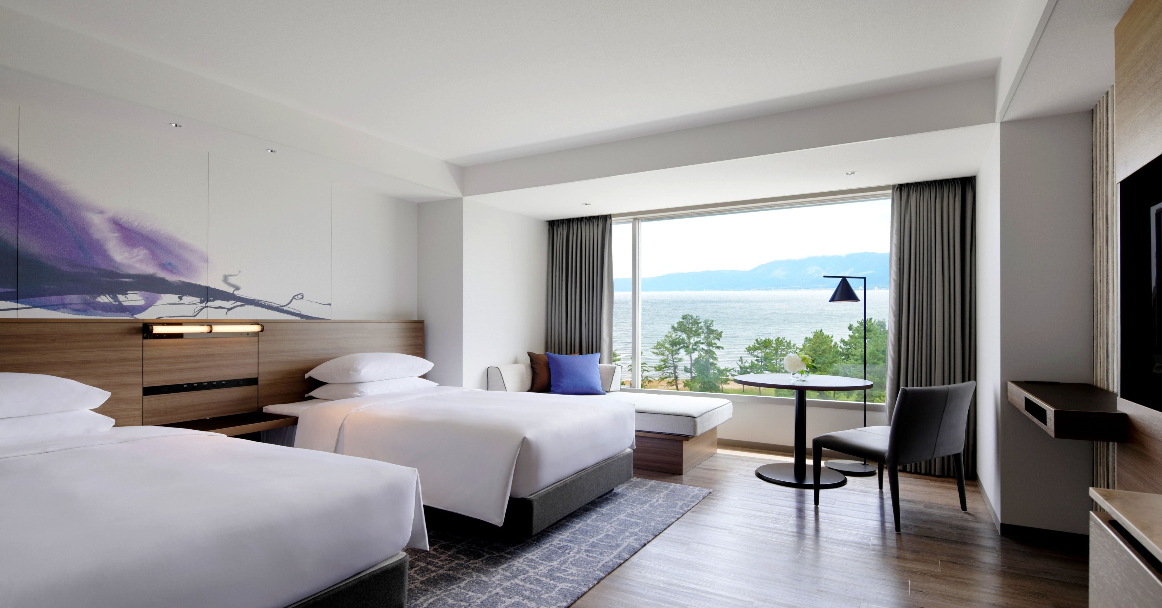 Een hotelkamer met twee bedden, een kleine tafel, een stoel en een groot raam met uitzicht op zee.