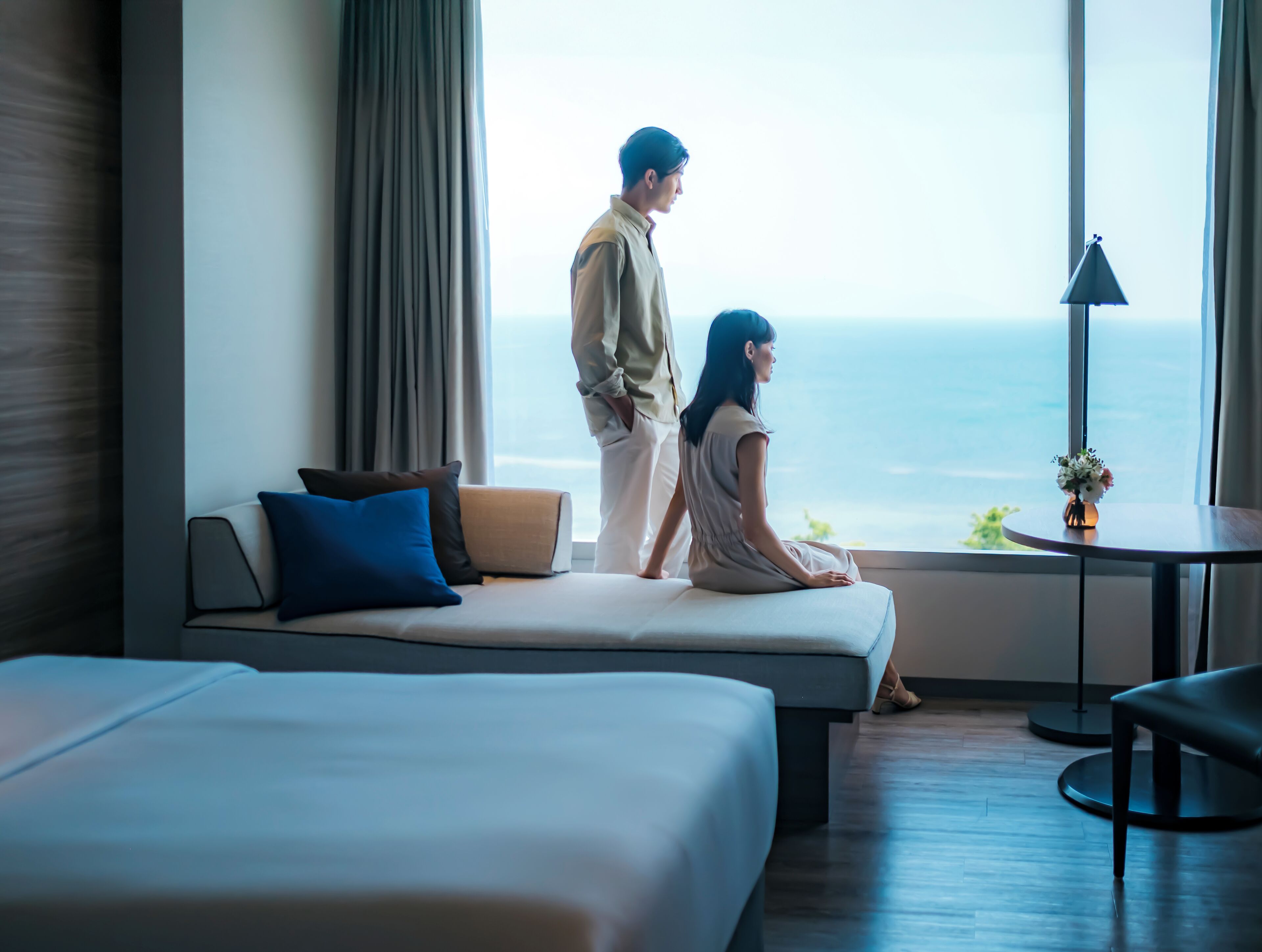 Habitación de hotel con cama, sofá, una mesita de noche con lámpara y vistas al mar.