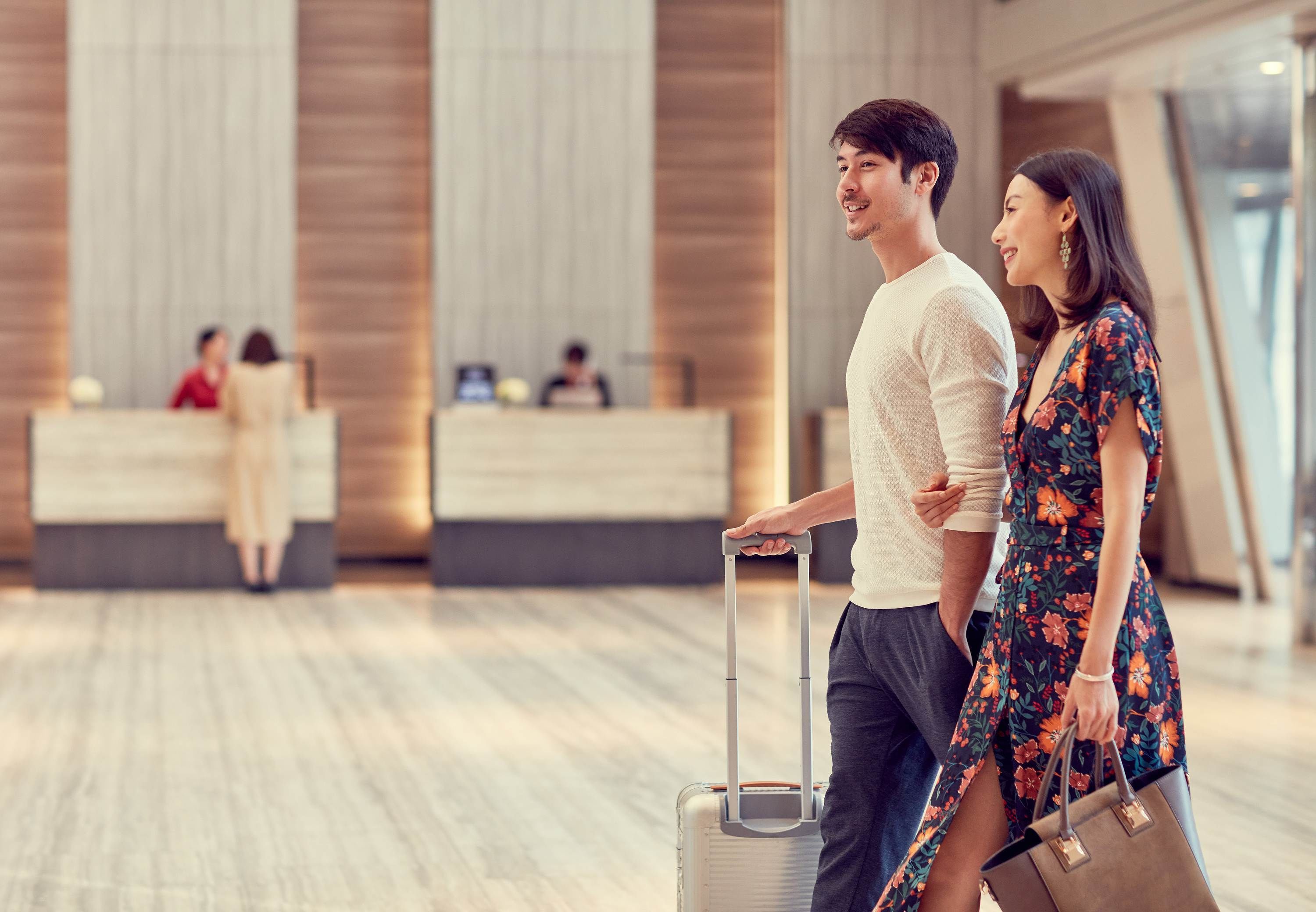 Um lobby moderno de hotel com um balcão de recepção, um homem puxando uma mala e uma mulher carregando uma bolsa.