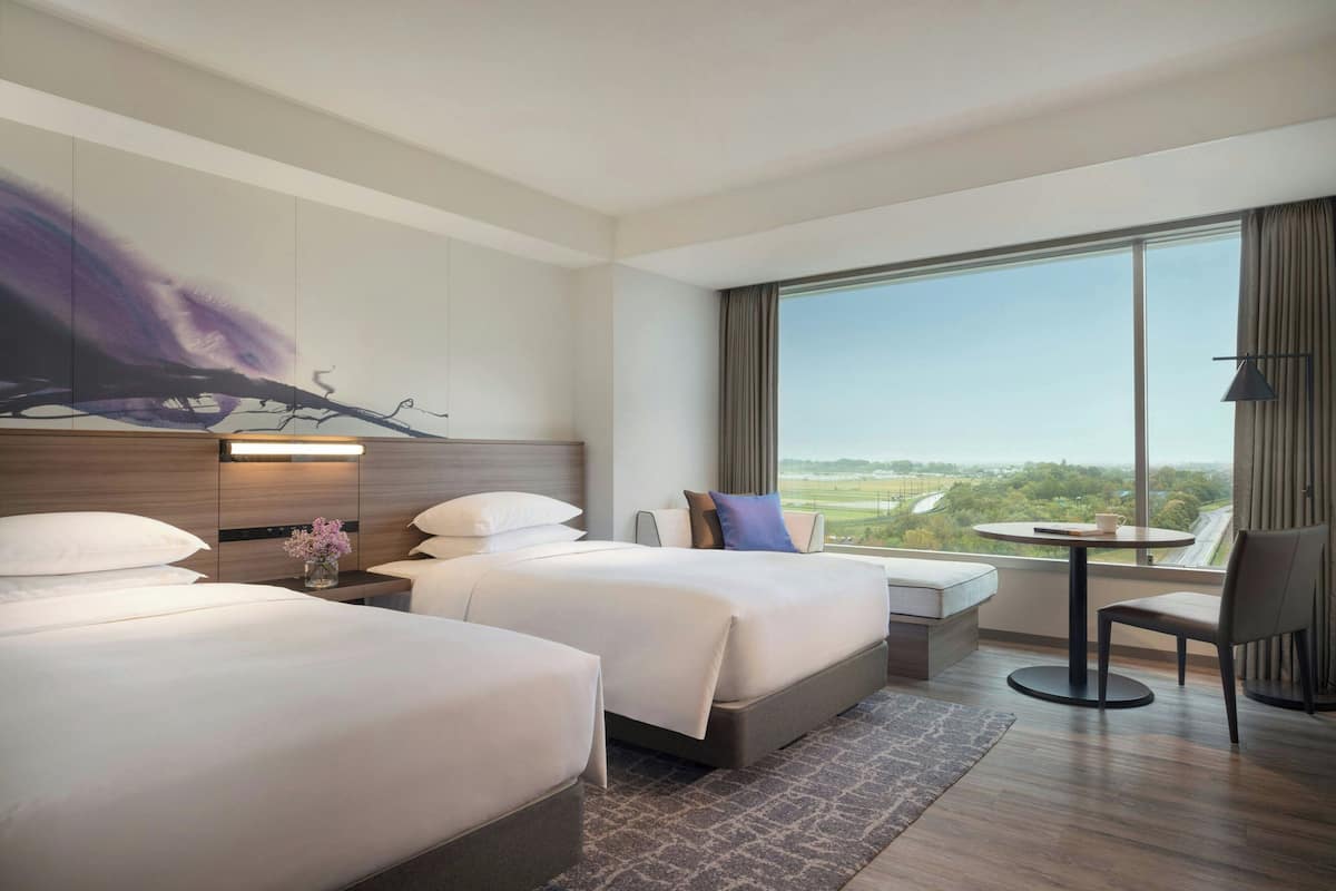 Une chambre d’hôtel avec deux lits, une petite table, une chaise et une grande fenêtre donnant sur l’extérieur.