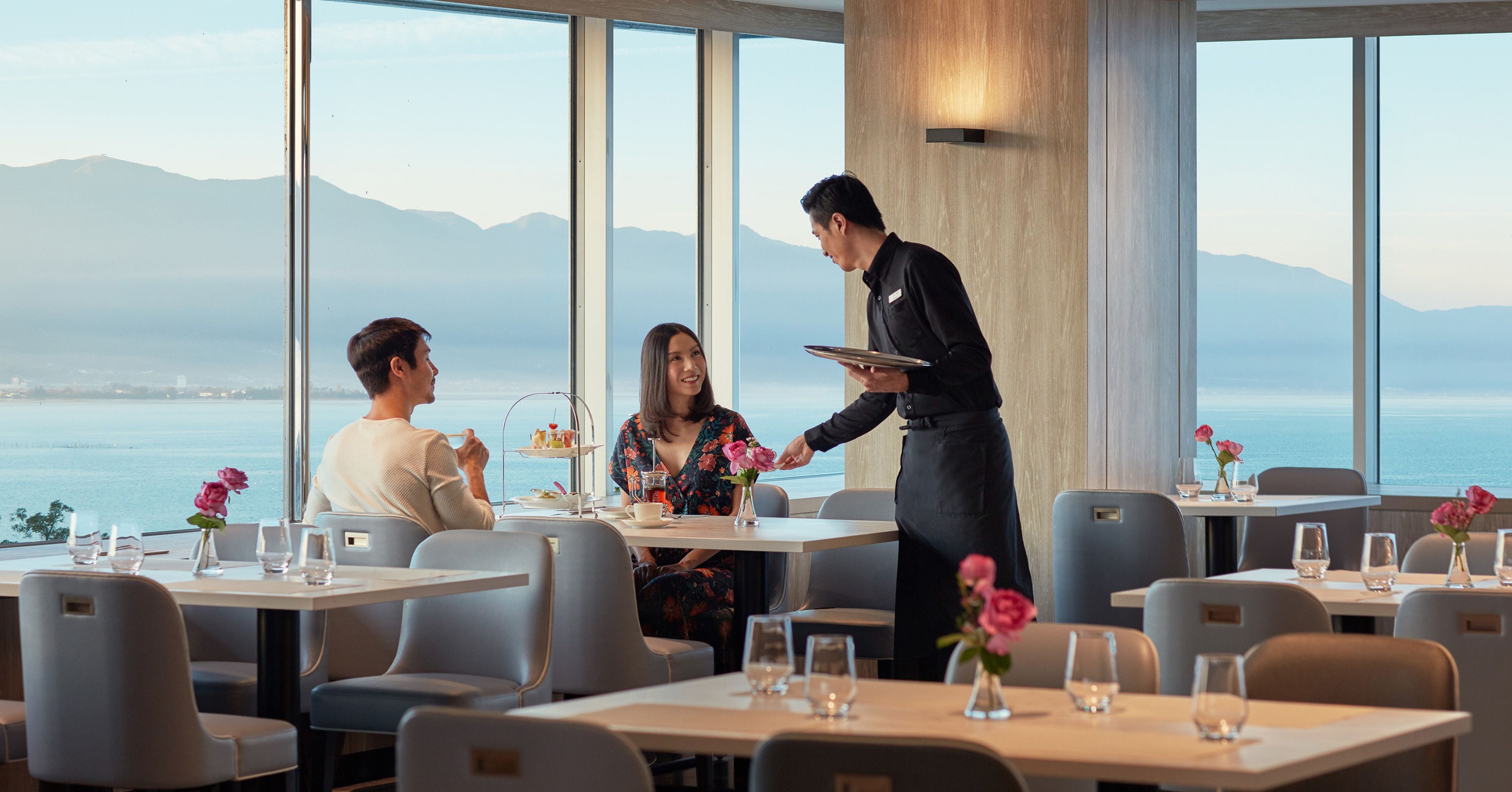 A restaurant with a view of the sea and mountains, tables with chairs, and a waiter serving a guest.