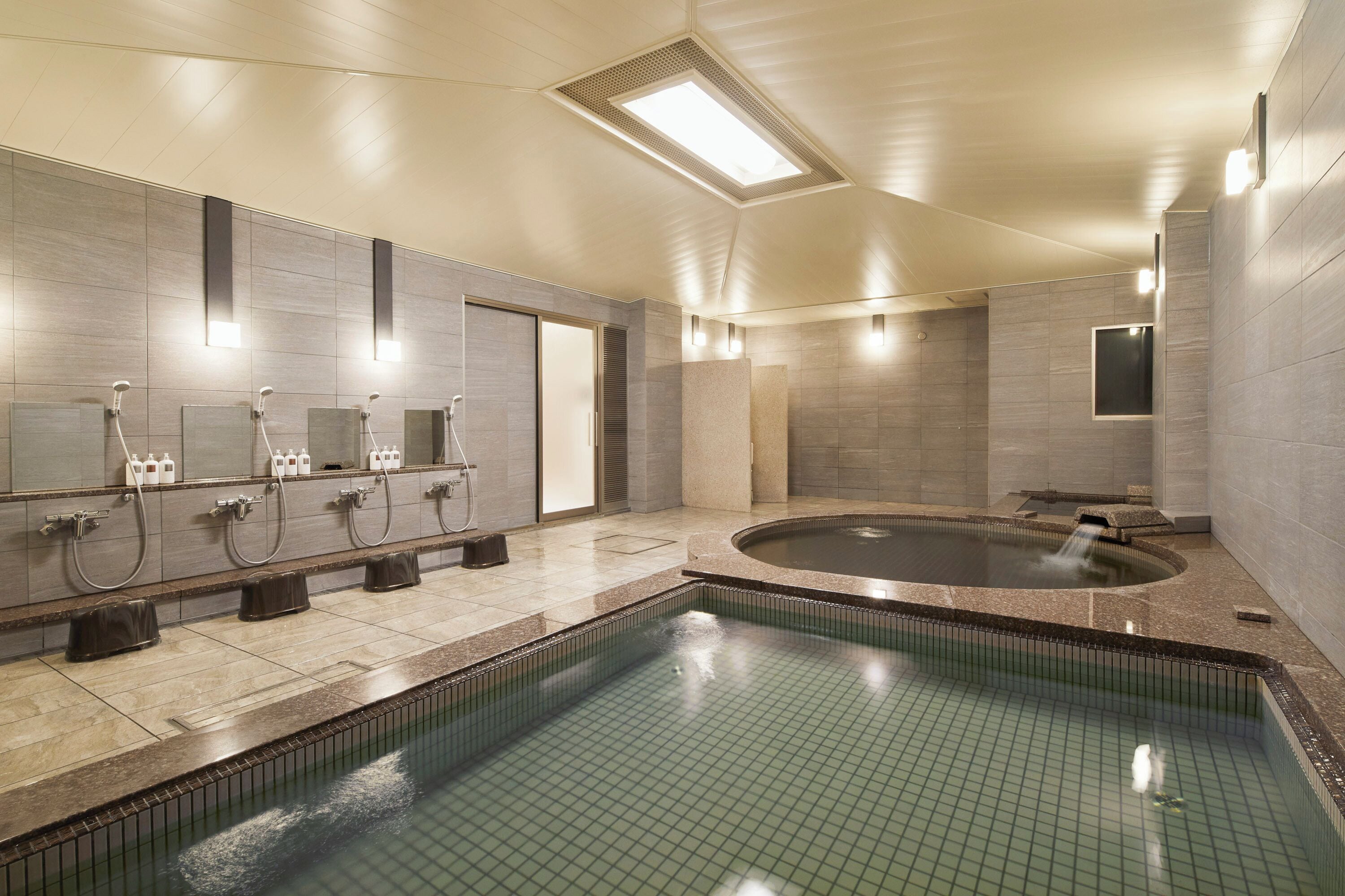 A modern indoor pool area with a rectangular pool, multiple shower stations, and a large hot tub.