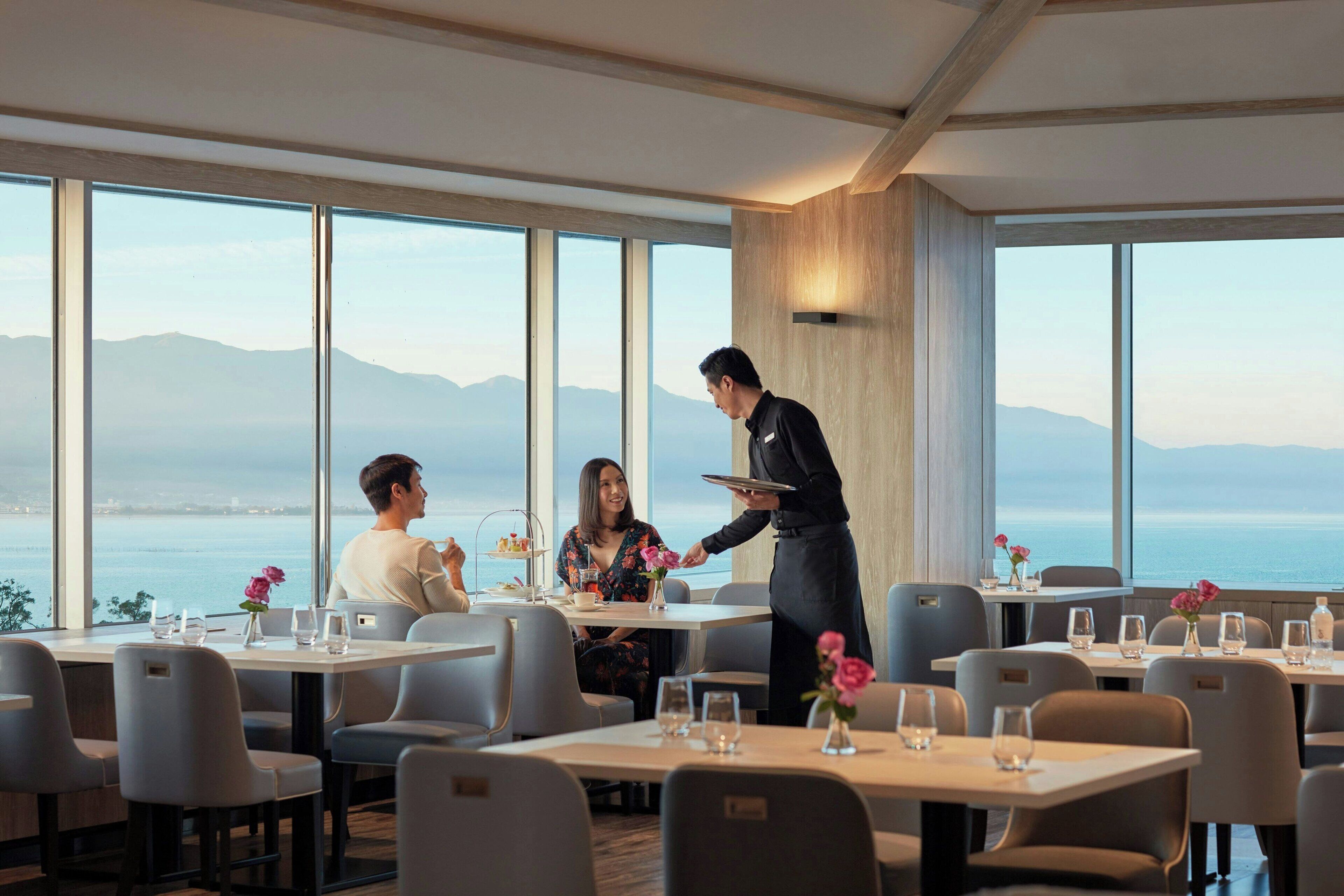 A restaurant with a view of the sea and mountains, featuring modern furniture and a waiter serving a guest.