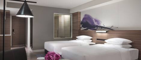 A modern hotel room with a large bed, a bedside table with a book and flowers, a desk with a chair, and a large mirror.