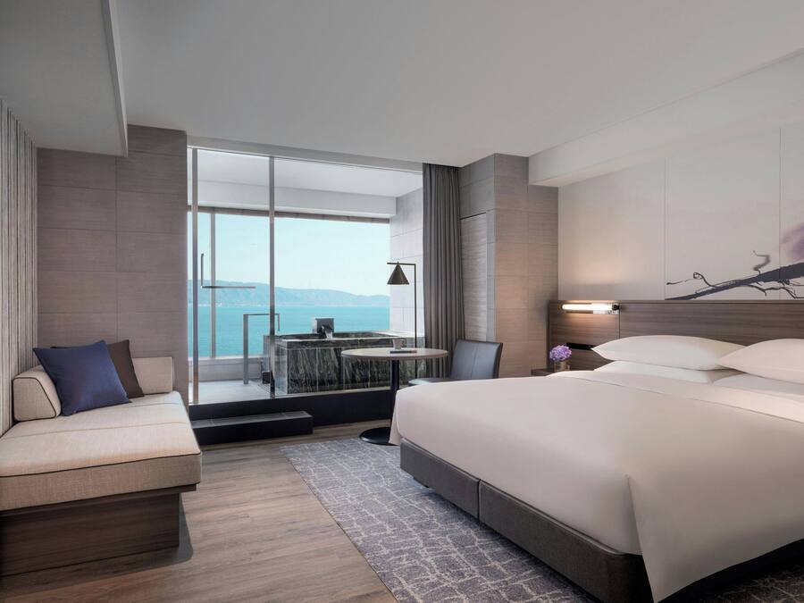 A modern hotel room with a large bed, a seating area with a view, and a small table with a lamp.