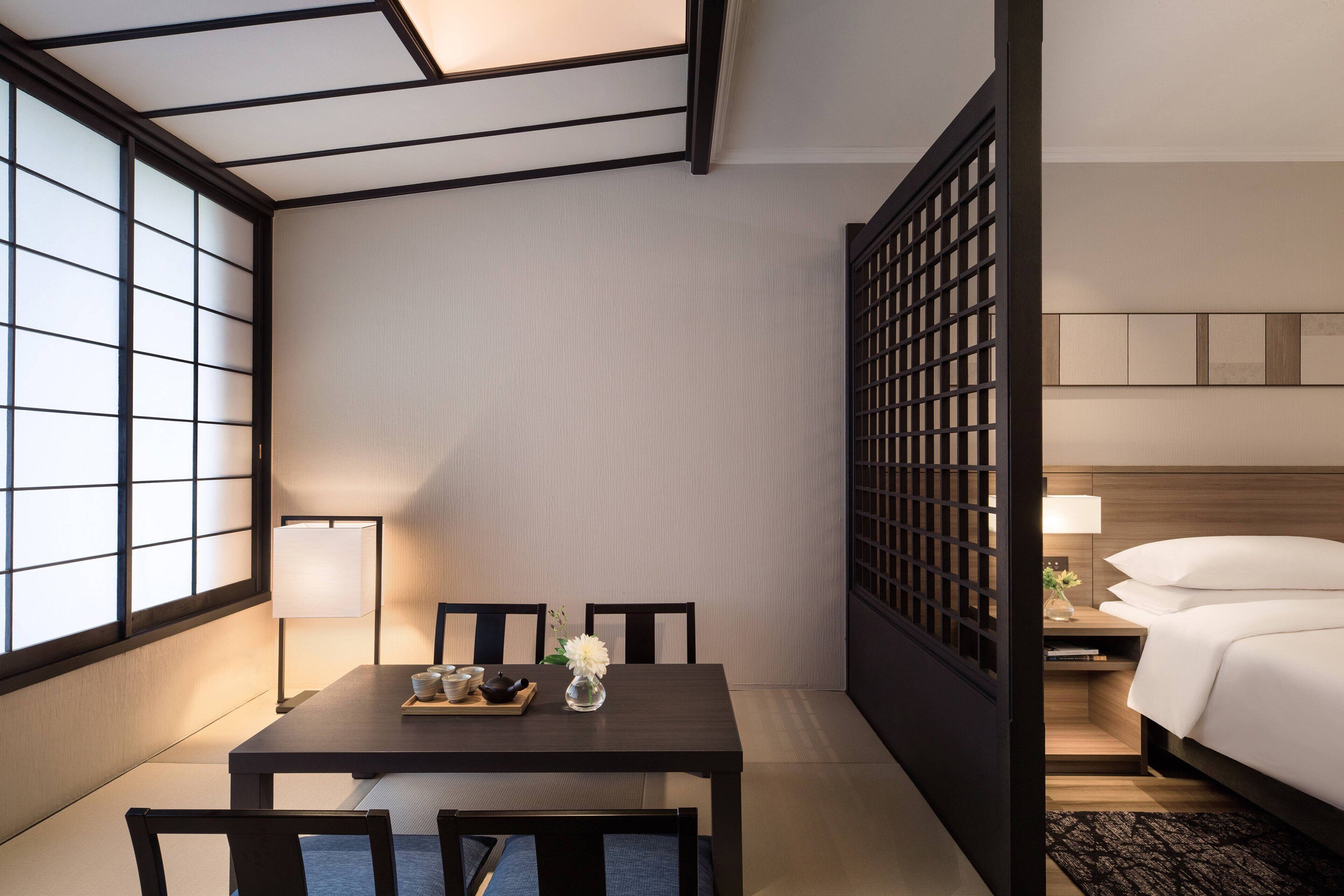 A hotel room with a tatami mat table, a bed with white bedding, a nightstand with a lamp, and a view through a shoji screen.