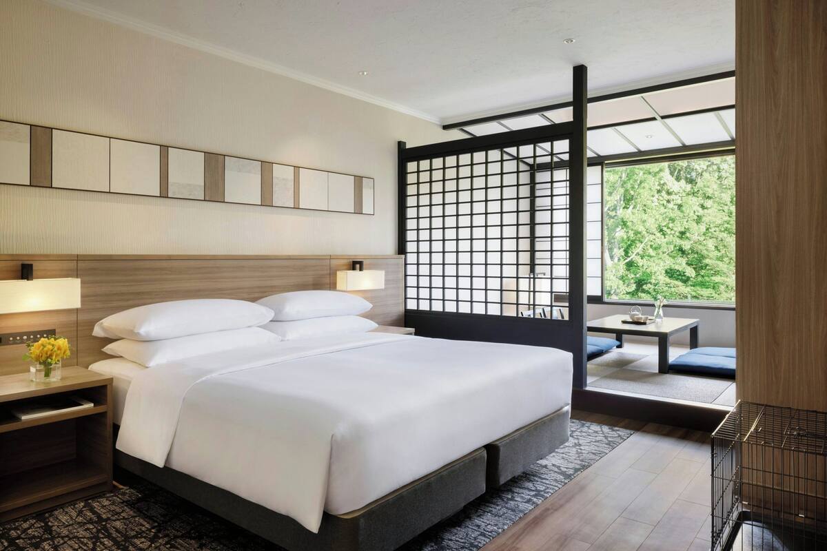 Ein modernes Hotelzimmer mit einem großen Bett, Nachttischen, einem kleinen Tisch und Blick auf Grün durch ein großes Fenster.