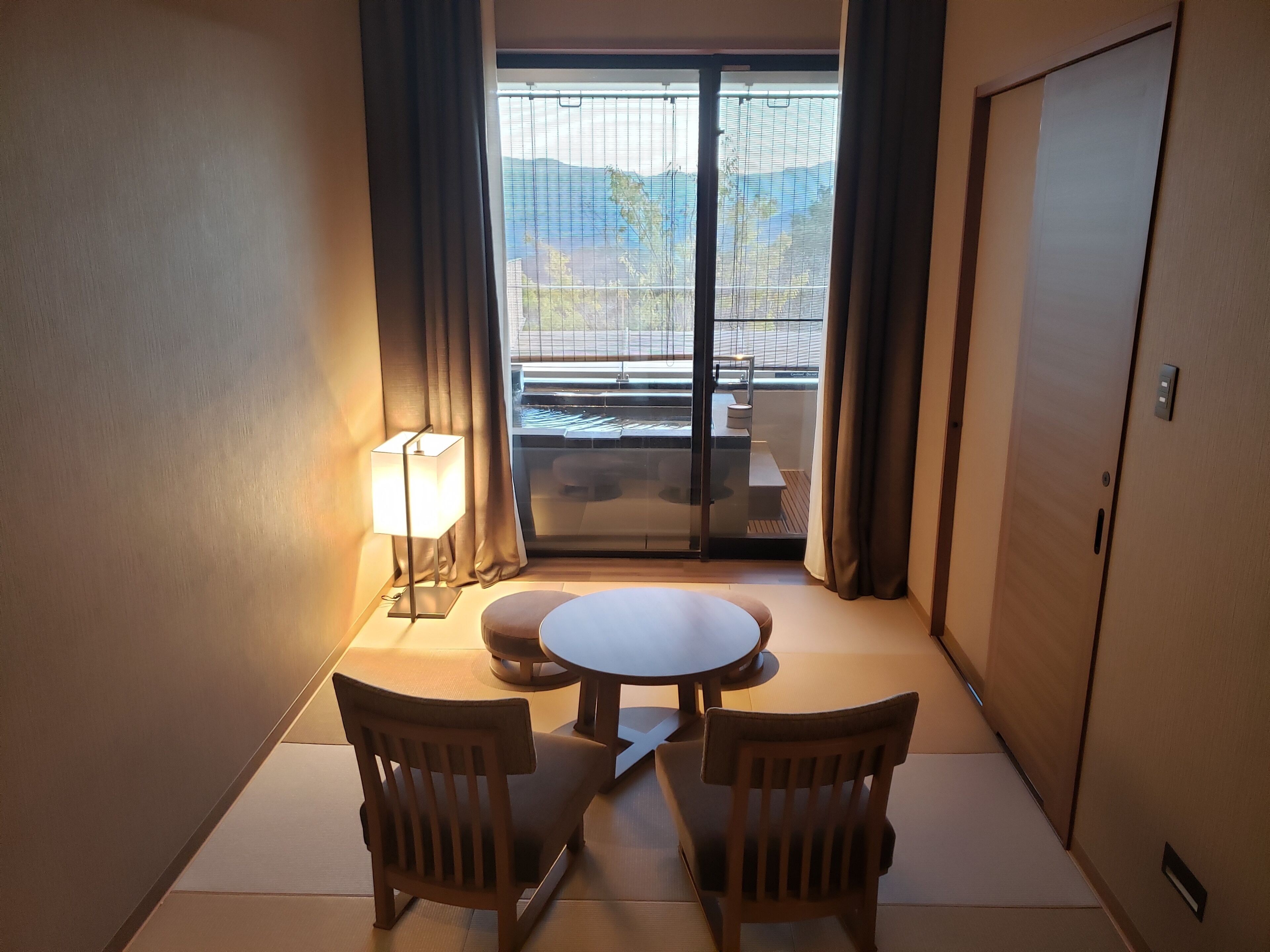 Habitación de hotel con una mesa redonda, dos sillas, una lámpara y un acceso corredizo a un balcón con vistas al exterior.