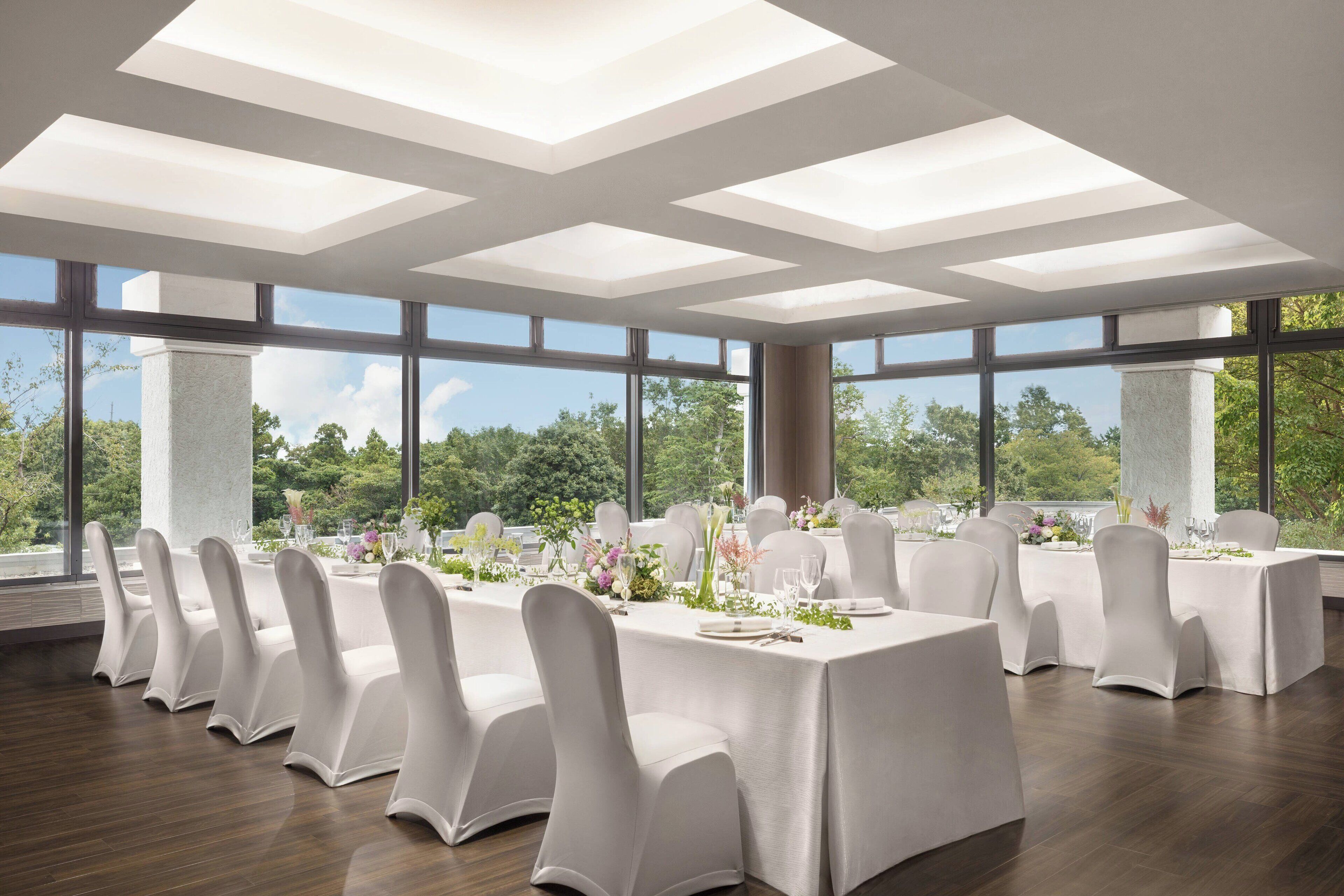 A spacious room with large windows, a high ceiling with skylights, and a long table set for a formal event.