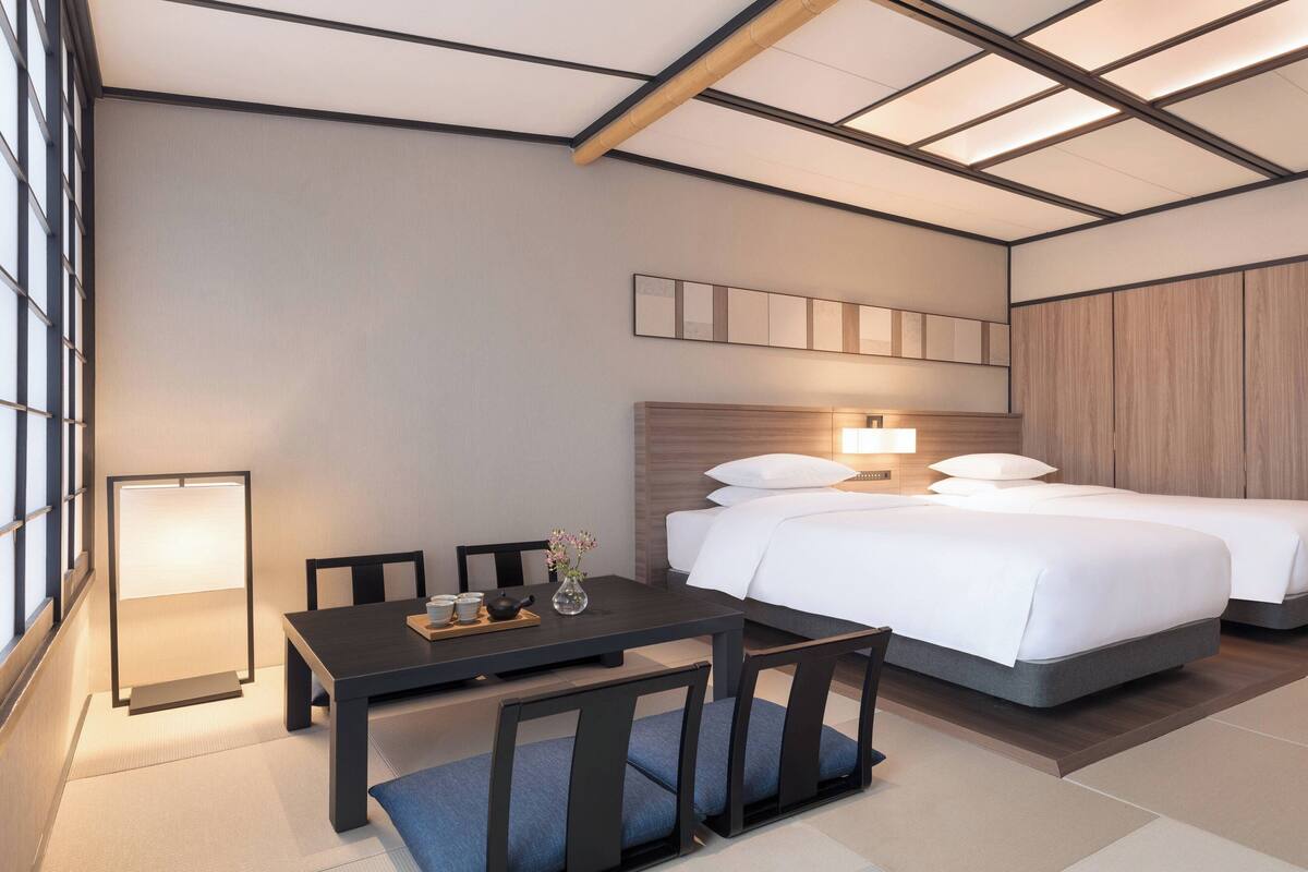 A hotel room with a bed, bedside table, lamp, and a view of the city.