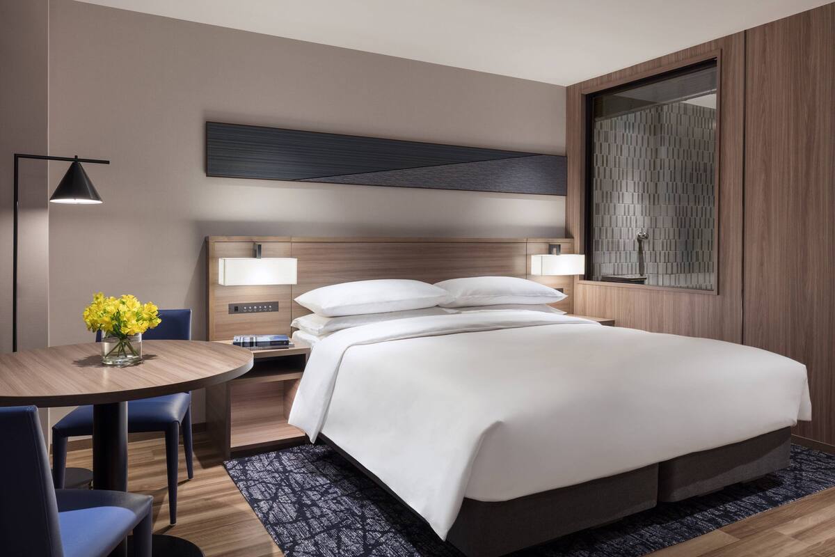 A modern hotel room with a large bed, bedside tables, a desk with a chair, a round table with chairs, and a vase of flowers.
