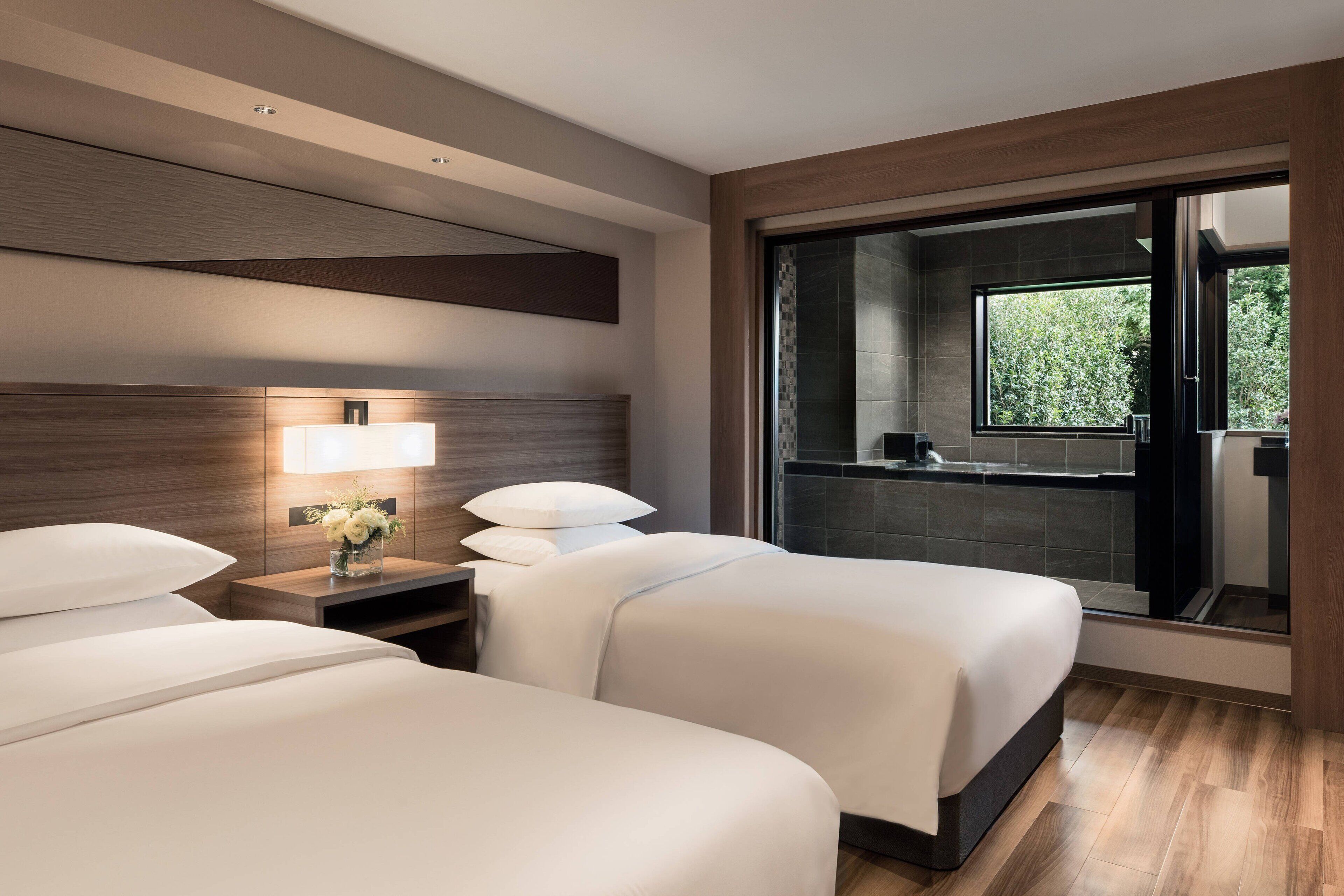 Habitación de hotel con dos camas, una mesita de noche con lámpara, una ventana con vistas a zonas verdes y una puerta corredera que da al baño.