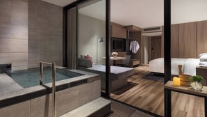 Een moderne hotelkamer met een glazen jacuzzi, een bed, een klein tafeltje met een vaas en een kaars, en een houten vloer.