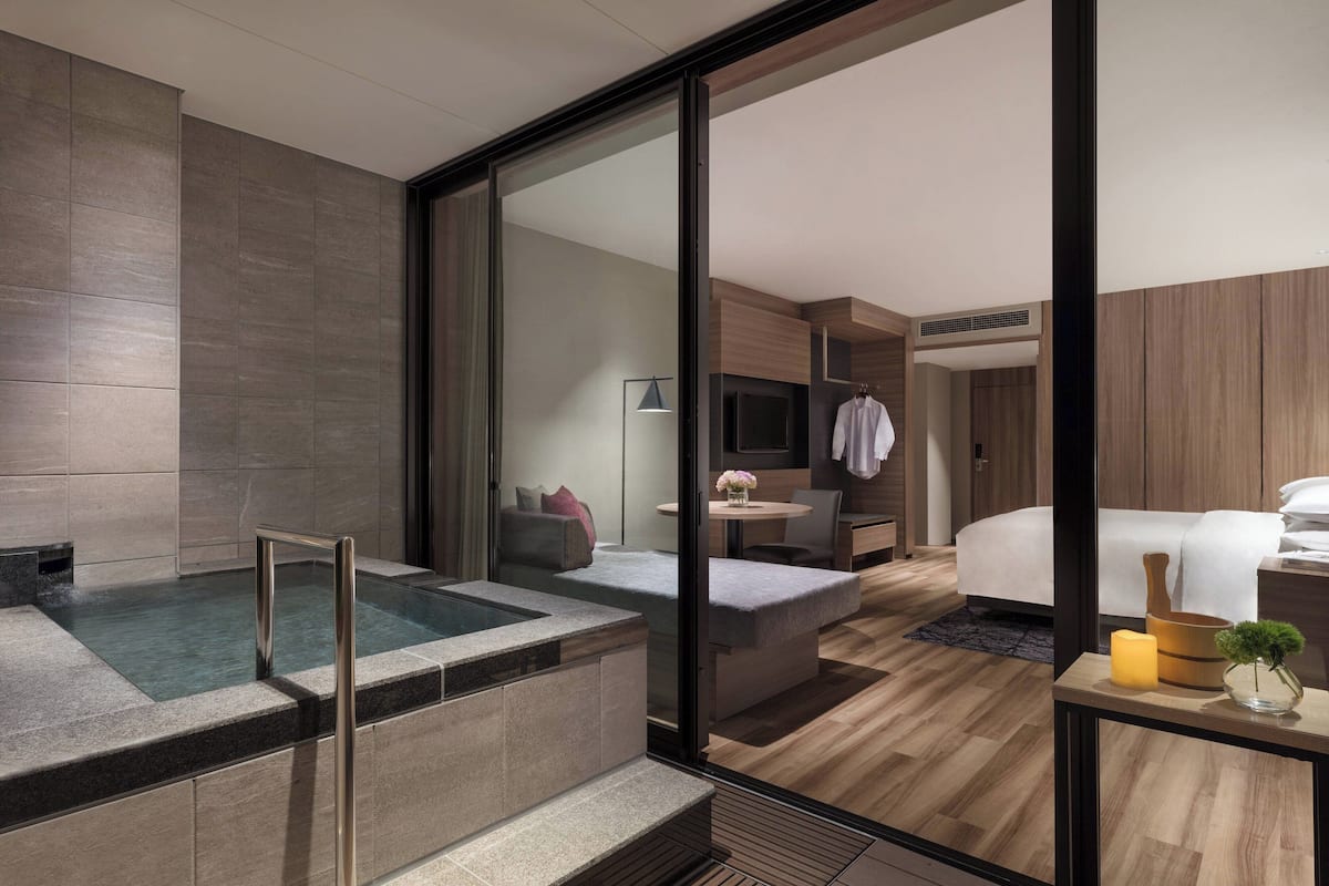 A modern hotel room with a glass-enclosed jacuzzi, a bed, a small table with a vase and candle, and a wooden floor.