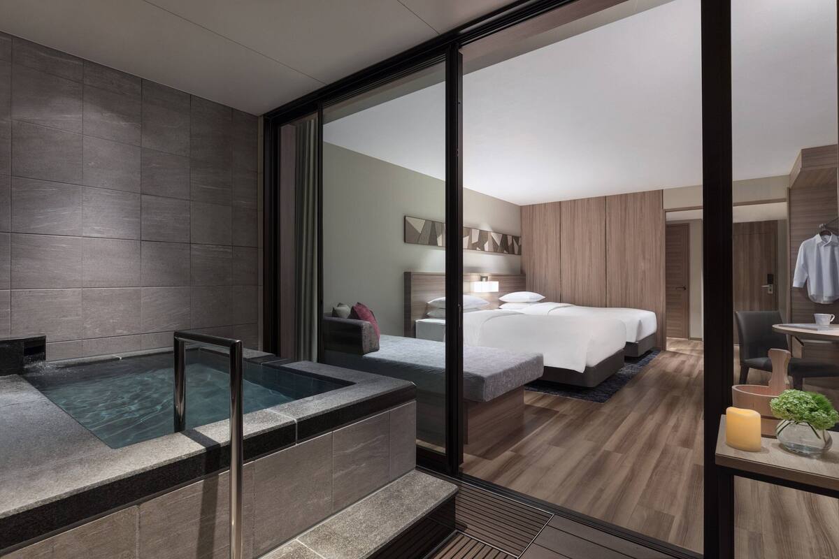 A modern hotel room with a glass-enclosed jacuzzi, a large bed, a bedside table with a candle, and a view into the adjacent room.