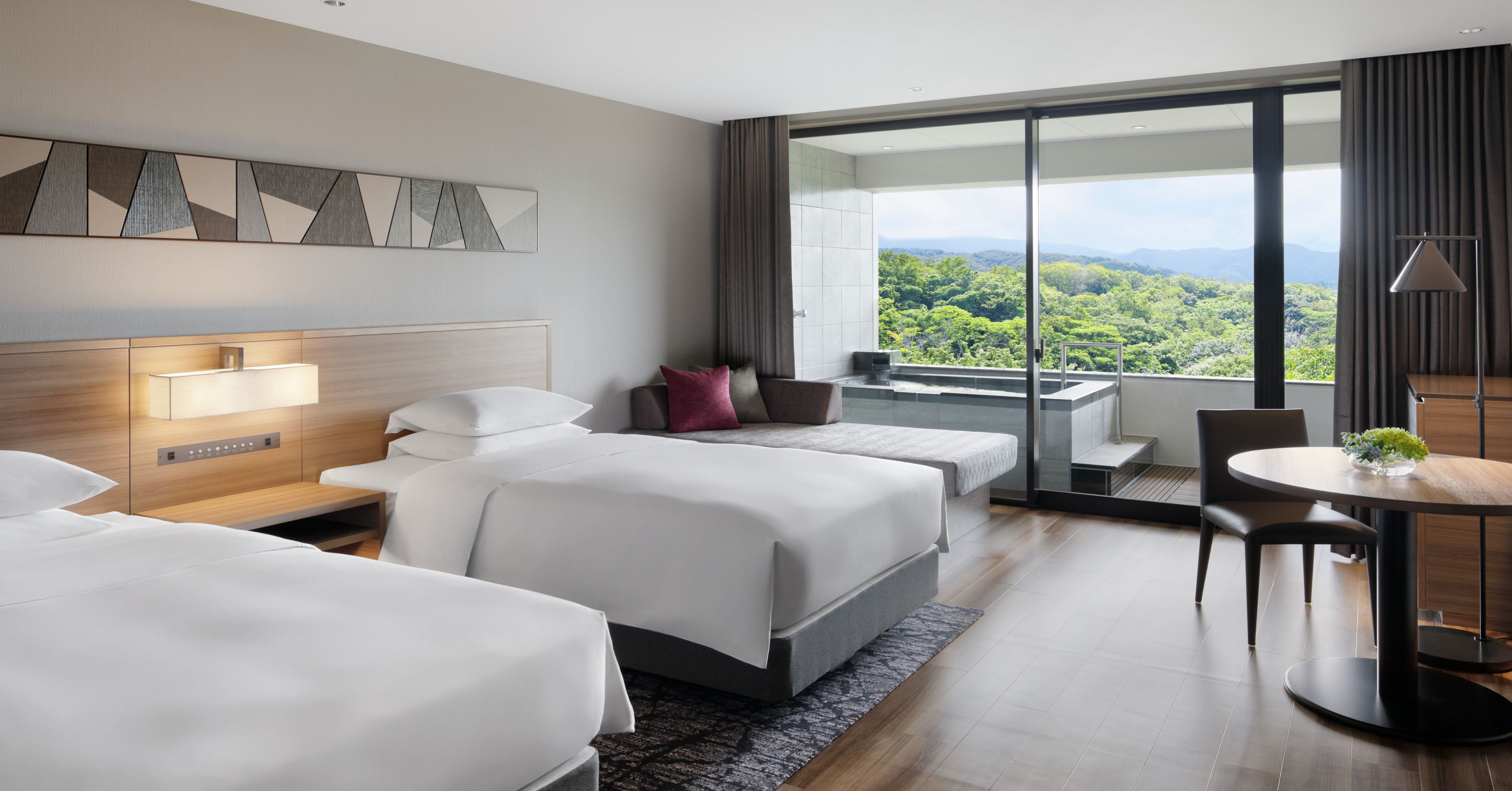 Habitación de hotel con dos camas, un escritorio, una silla, una mesita con una planta y un ventanal con vistas al exterior.