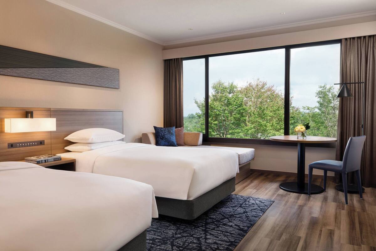 A hotel room with two beds, a small table with a vase, a chair, and a view of greenery through large windows.