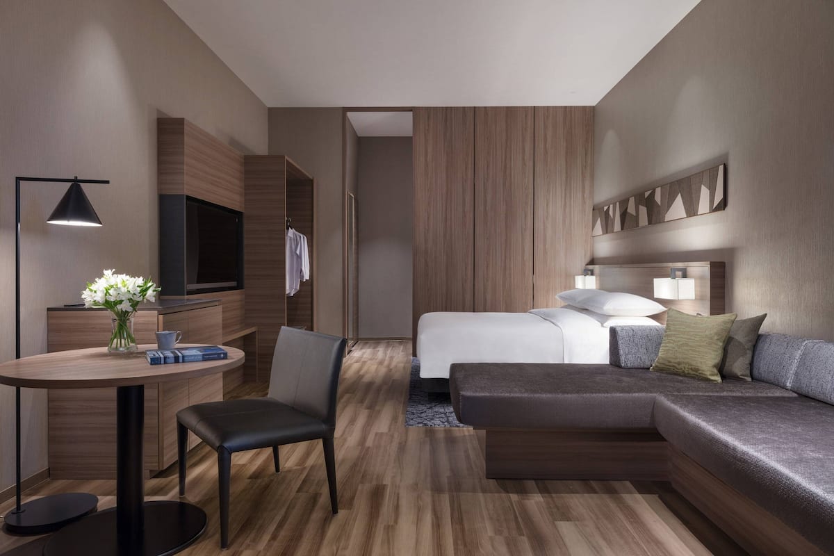 A modern hotel room with a wooden floor, a sofa bed, a desk with a chair, a TV, and a bedside table with a vase of flowers.