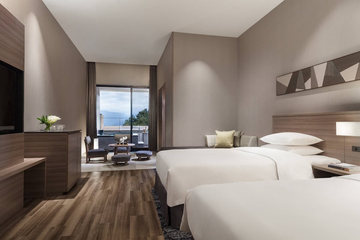A hotel room with two beds, a desk, a chair, a small table with a vase, and a view of the outdoors.