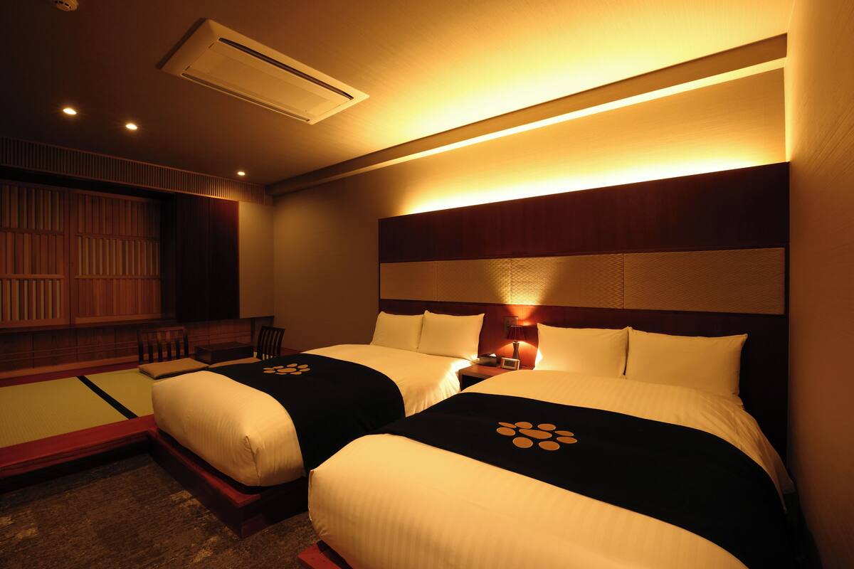 A hotel room with two beds, a wooden headboard, a bedside table with a lamp, and a futon area.