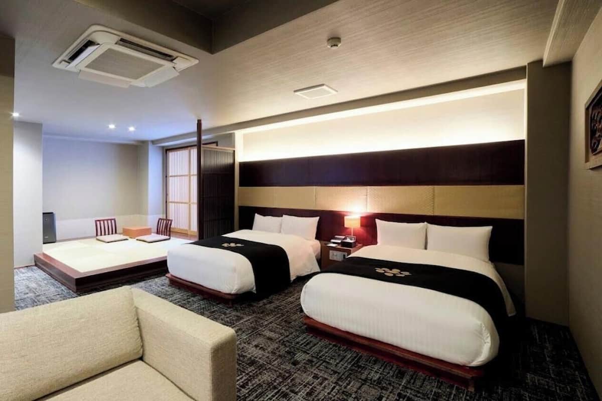 A hotel room with two beds, a sofa, a bedside table, and an air conditioning unit.