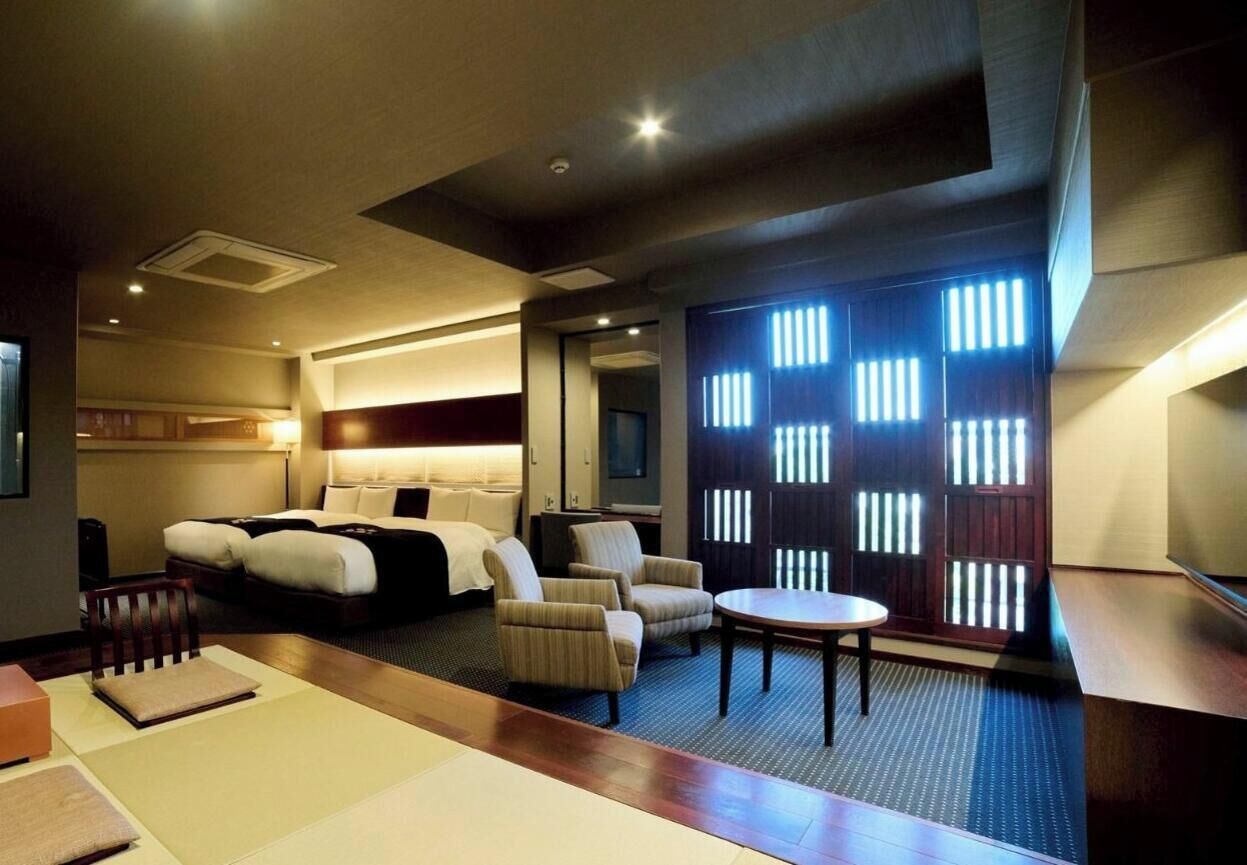 Una habitación de hotel con una cama, un área de descanso con sillas, una mesita y un separador con listones verticales.