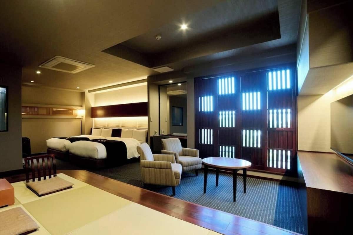A hotel room with a bed, a seating area with chairs, a small table, and a partition with vertical slats.