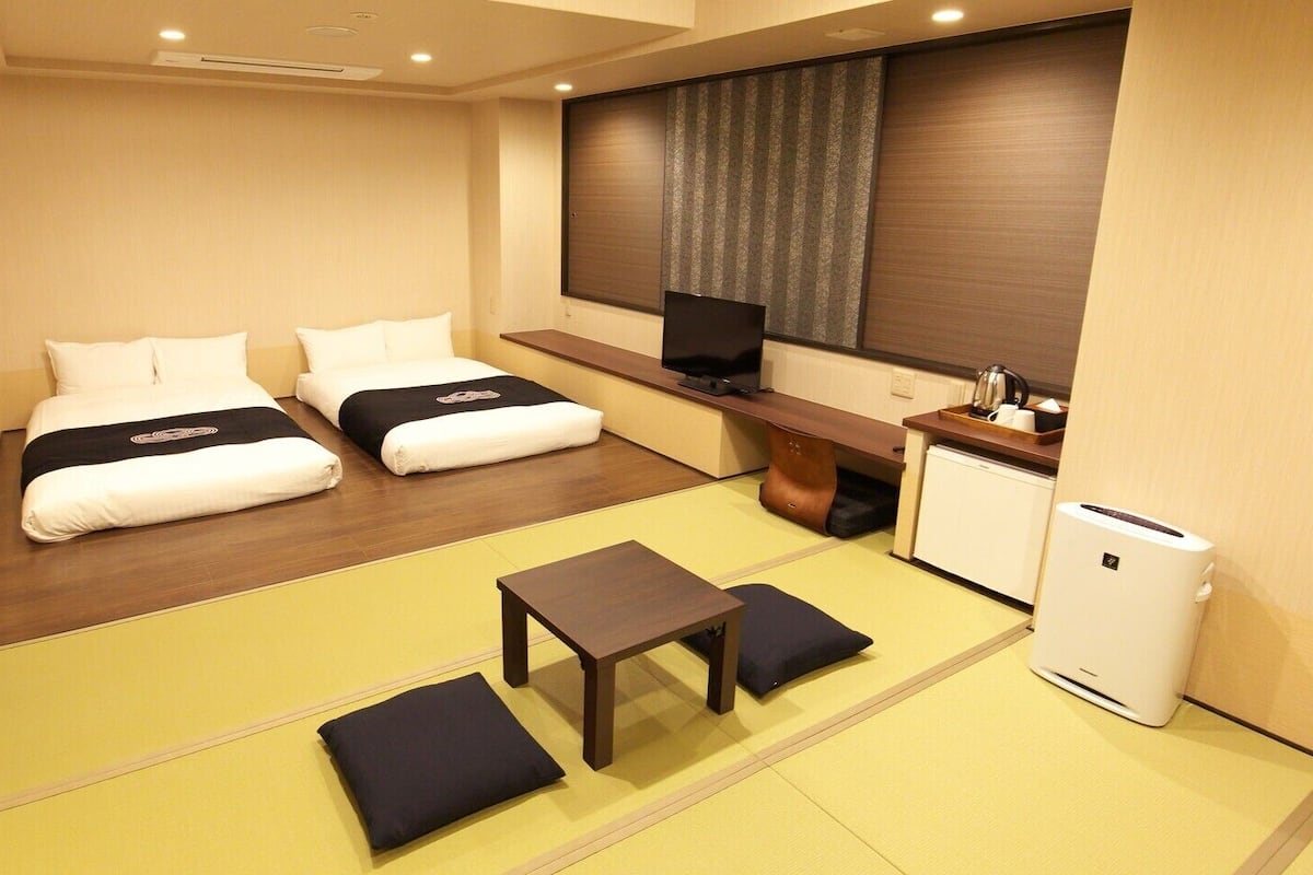 Quarto de hotel com duas camas, uma mesa pequena, televisão e cozinha compacta.