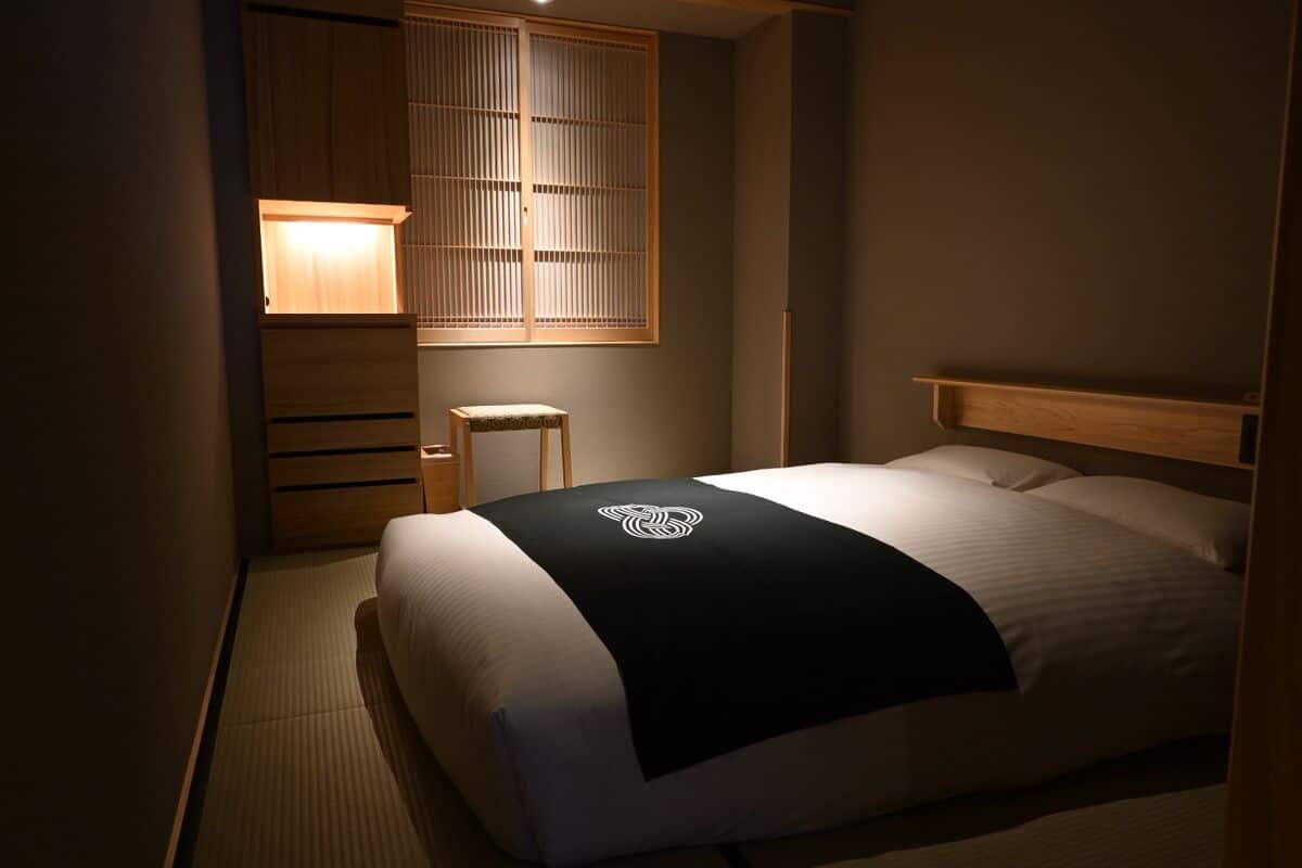A room with a bed, a small stool, a wooden cabinet, and a sliding door with a light.