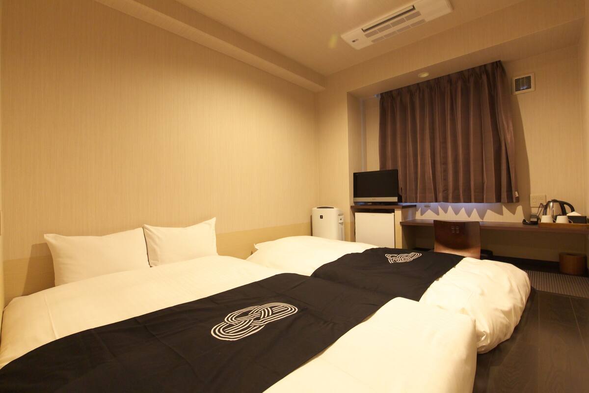 Quarto de hotel com duas camas, uma escrivaninha, uma cadeira, uma televisão e uma janela com cortinas.