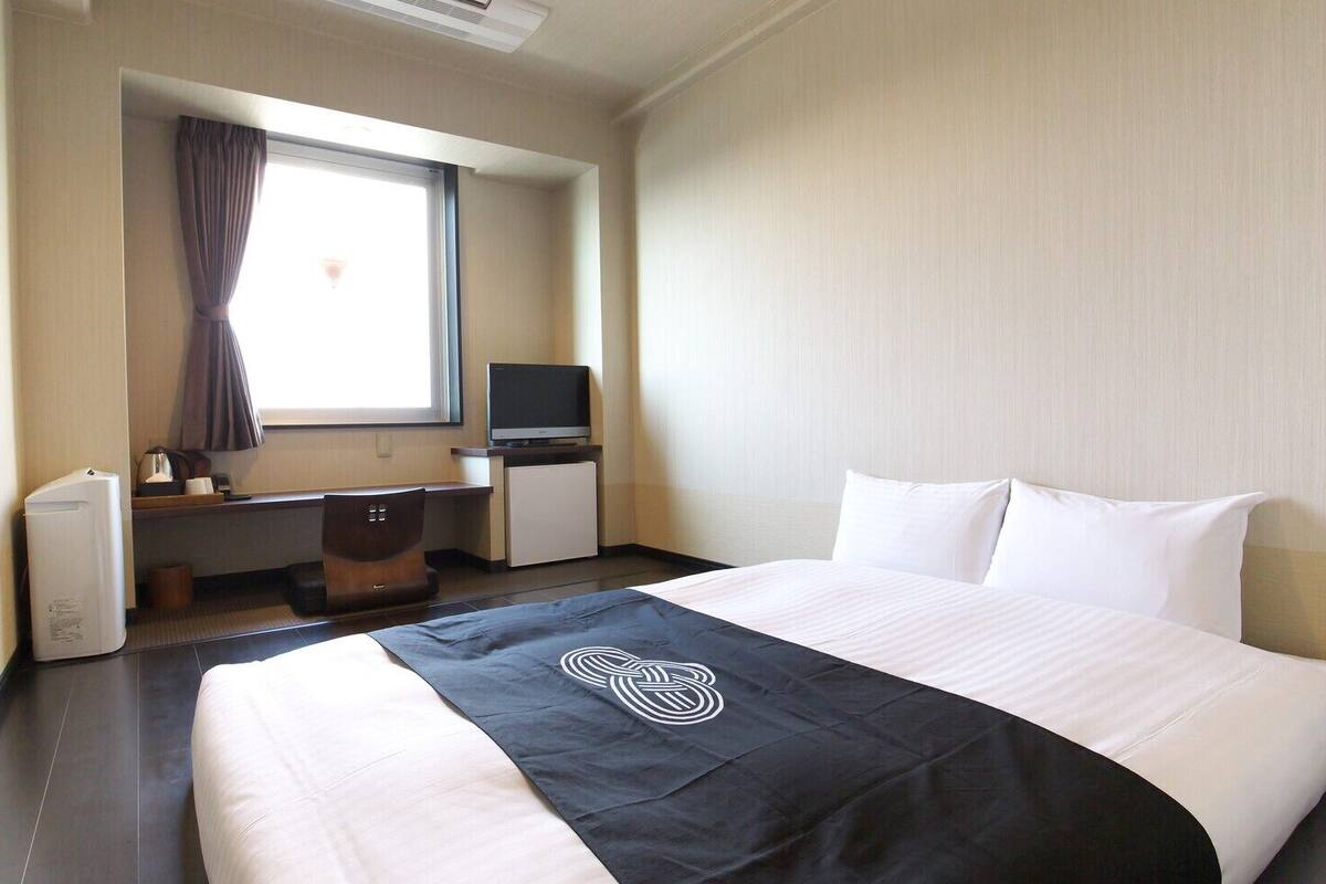 Quarto de hotel com cama, escrivaninha, televisão, mini-geladeira e janela com cortinas.