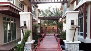 A red wooden bridge leading to a covered area with stone pillars and a view of a building with large windows. - Beachfront Family Penthouse Hua Hin (Hua Hin)