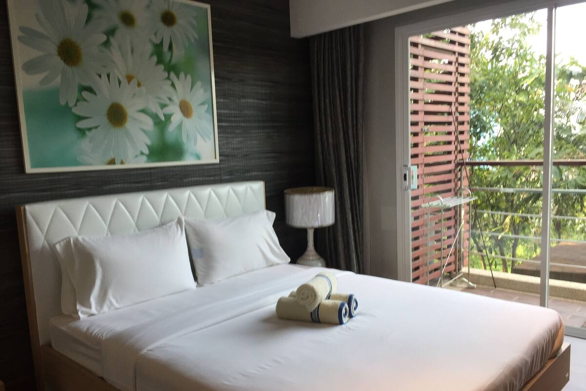 A hotel room with a large bed, a painting of daisies, a window with a view of greenery, and a nightstand with a lamp.
