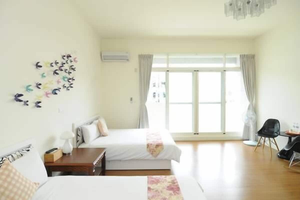 A hotel room with two beds, a desk, and a chair. There is a large window with curtains, and a wall decoration of small figures.