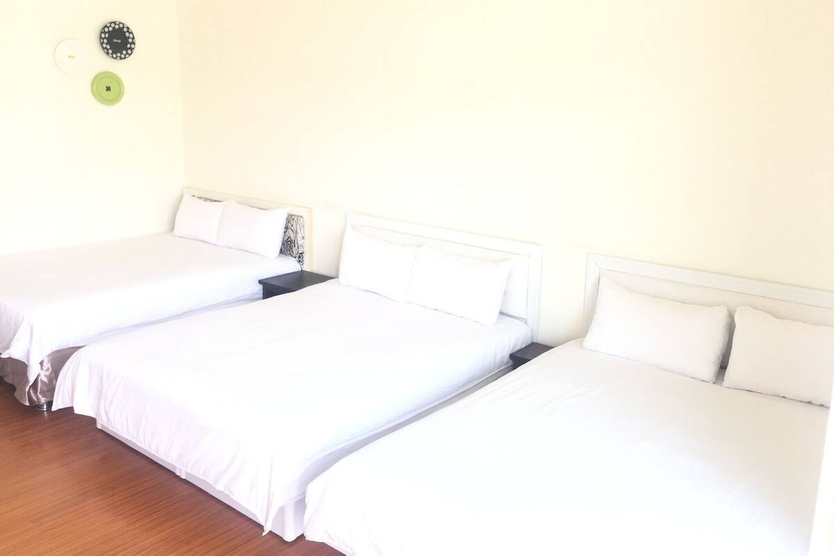 A hotel room with three single beds, each with white bedding and headboards. There is a small table between the beds, and a wall-mounted clock above it.