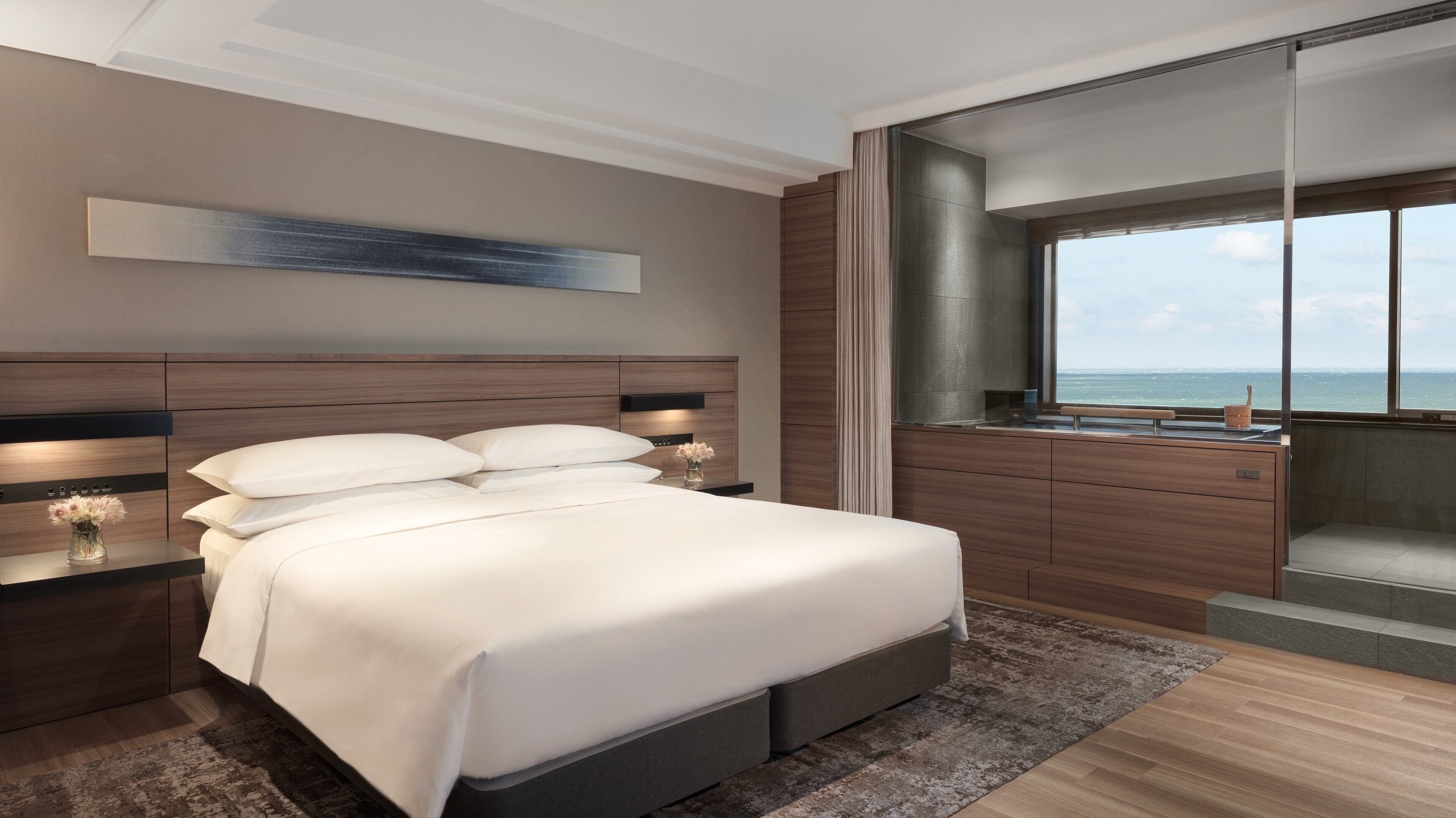 A modern bedroom with a large bed, a nightstand with a vase of flowers, a dresser, and a view of the ocean through a large window.