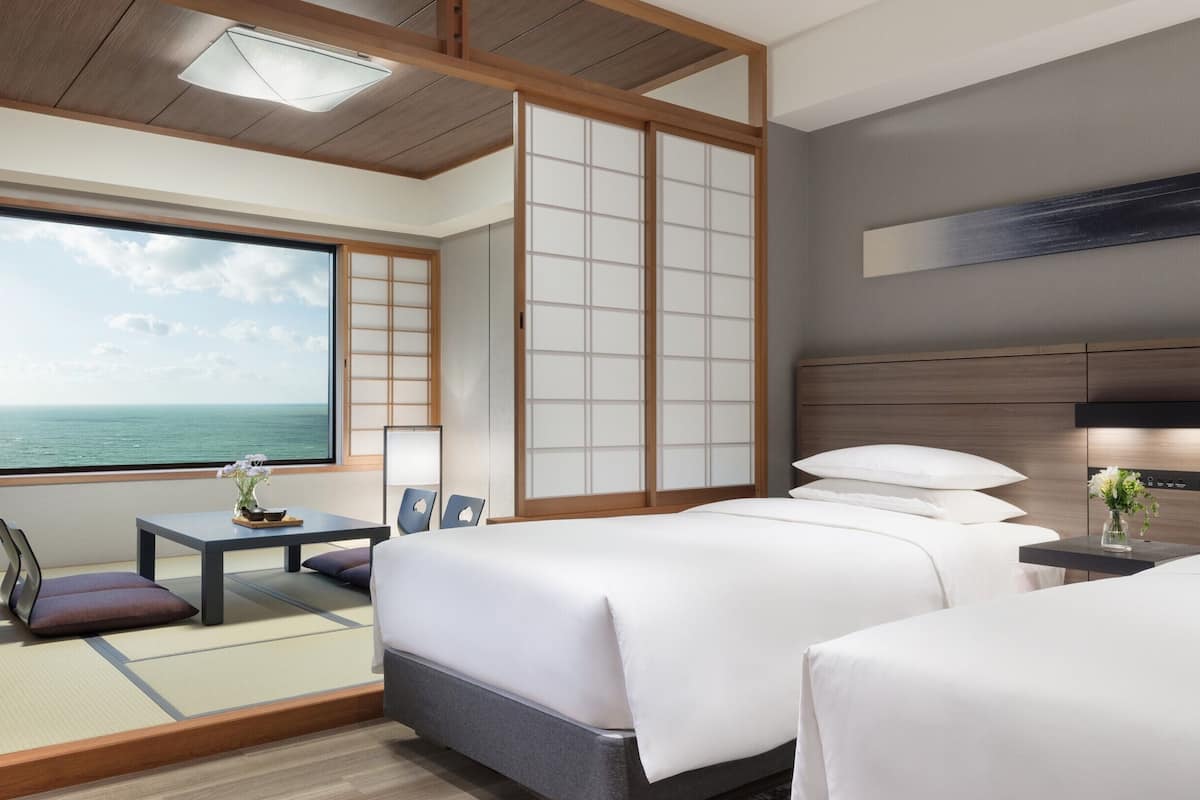Een hotelkamer met een groot raam met uitzicht op zee, twee bedden met witte lakens, een klein tafeltje met een bloevas, en een traditionele Japanse schuifdeur.
