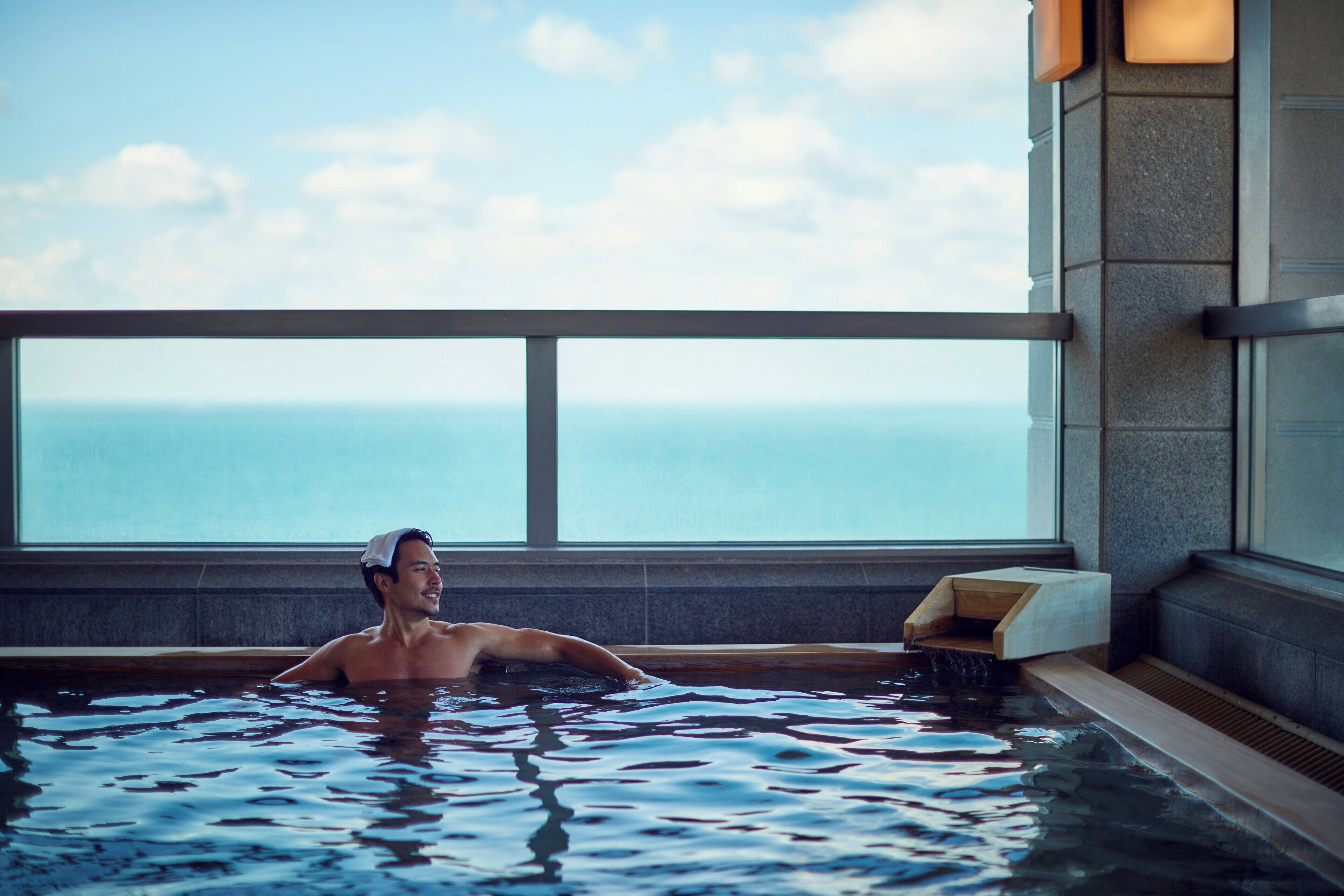 A person in a hot tub with a view of the ocean through large windows.