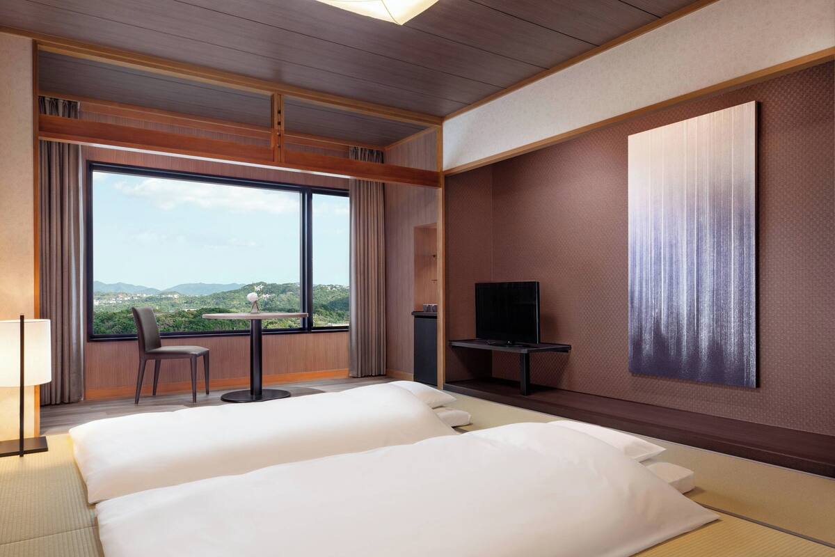 Een hotelkamer met twee bedden, een televisie, een tafel en uitzicht op groen door grote ramen.