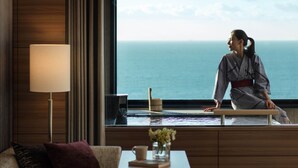 Ein Zimmer mit Meerblick, ein Sofa, ein kleiner Tisch mit einer Lampe und eine Person, die am Fenster sitzt.