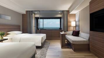 A modern hotel room with two beds, a sofa, a TV, and a large window with a view of the sea.