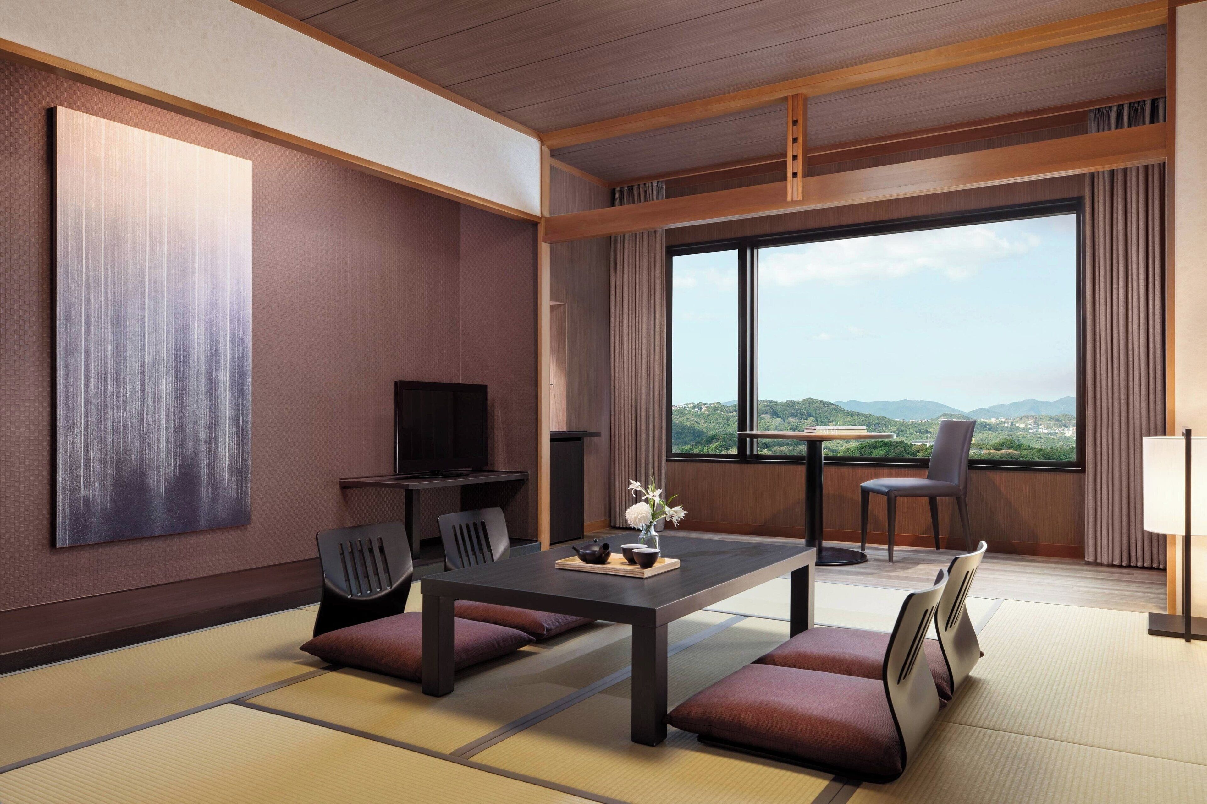 Une chambre d’hôtel moderne avec un sol recouvert de tatamis, une grande fenêtre donnant sur le paysage, un téléviseur à écran plat et un service à thé japonais traditionnel posé sur la table.