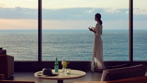 Ein Zimmer mit großem Fenster zum Meer, einem runden Tisch mit Flasche, einer Blumenvase und einem Hut.