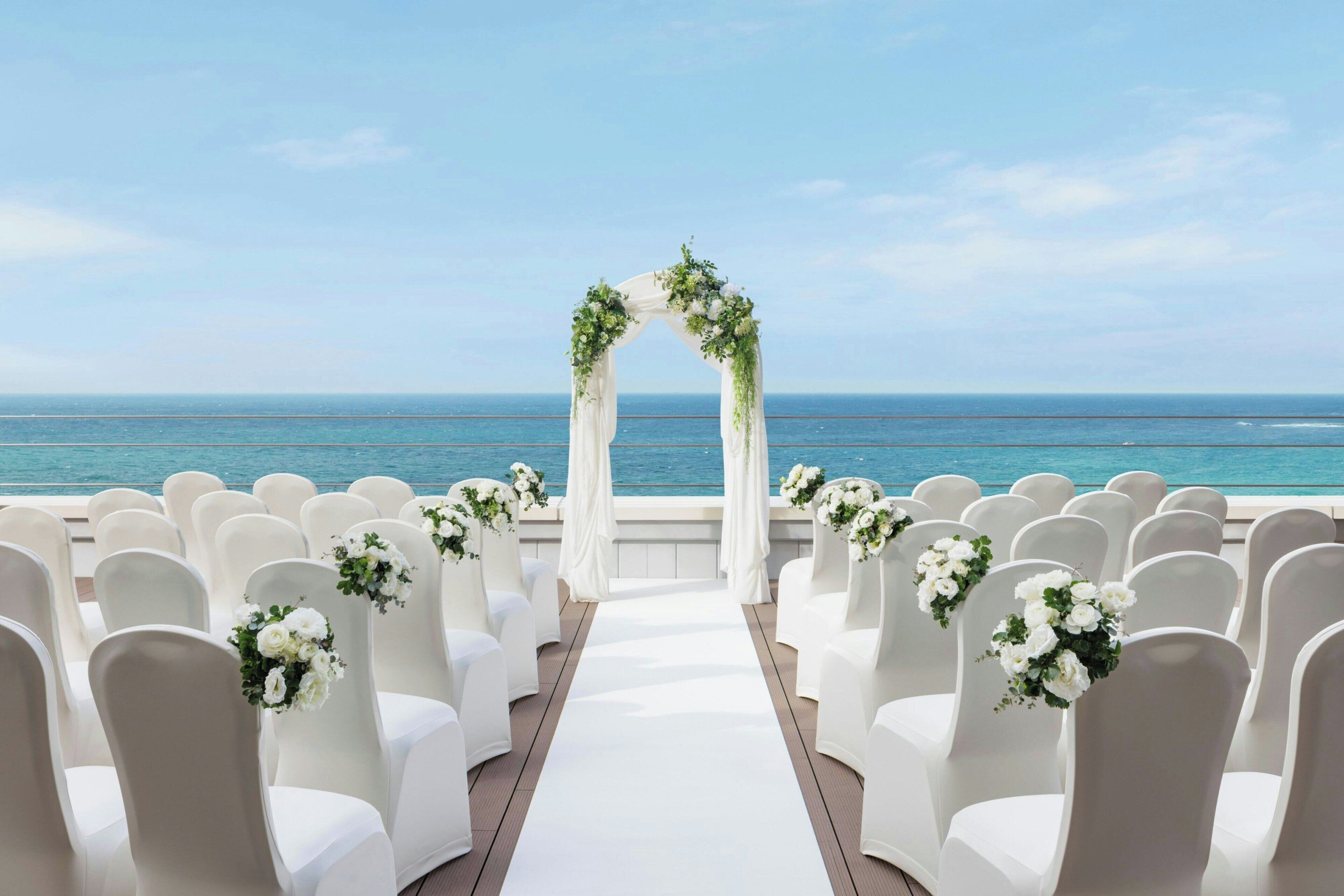 A wedding aisle with white chairs and a floral arch, leading to a view of the ocean.